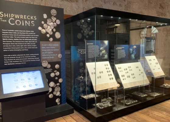 A cabinet displaying silver coins in many currencies, found on shipwrecks from the Western Australian coast. The coins glimmer in the cabinet spotlight. Next to the showcase is an information panel and a touch screen featuring more information on the coins.