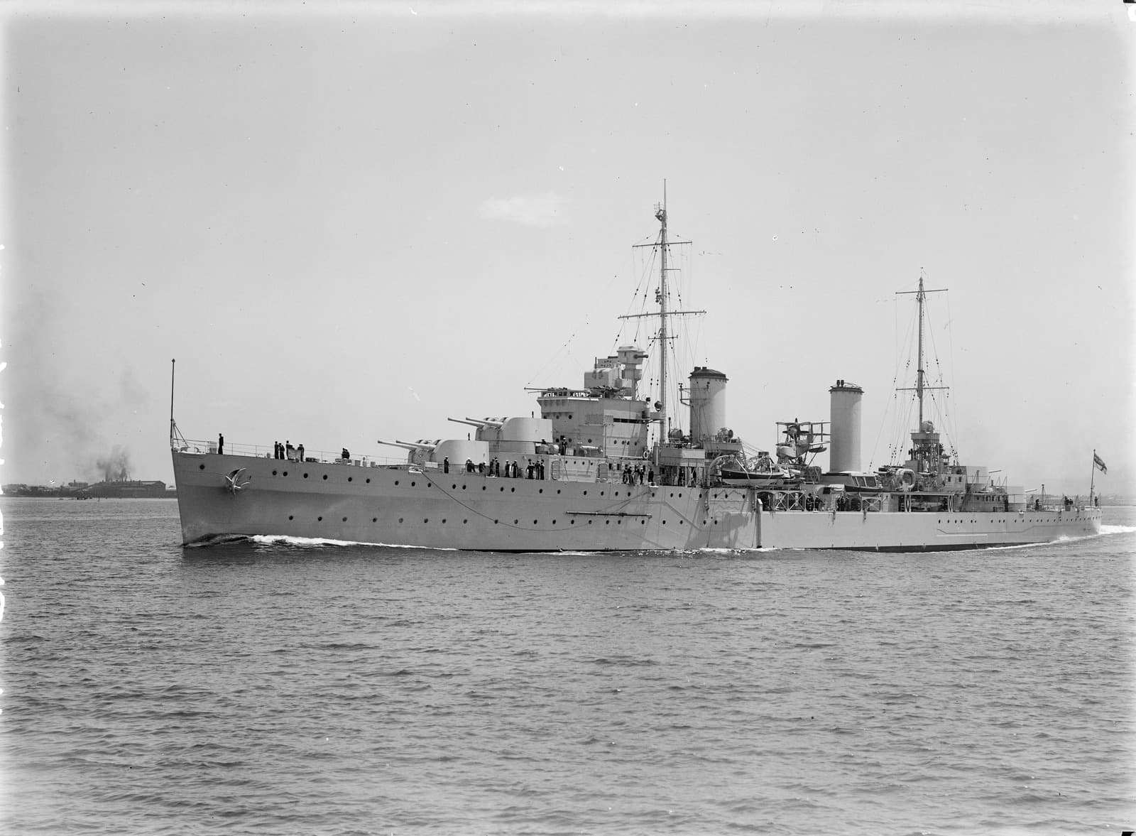 Naval ship with two masts and two turrets. Large guns and many sailors are on the deck of the ship. The ship is sailing away from land in the distance.