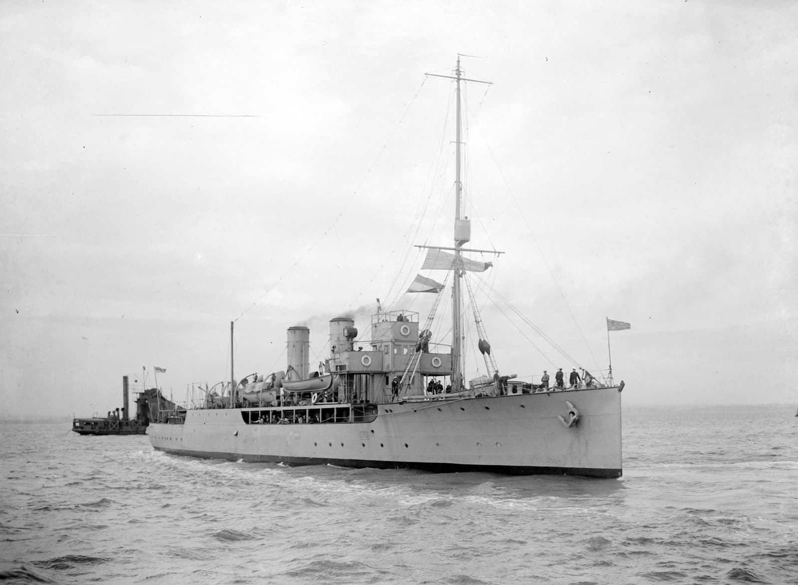 Naval style ship with a mast, two turrets and a bridge. The ship is flying a few flags and people are visible on the deck. There is a smaller ship in the distance behind.