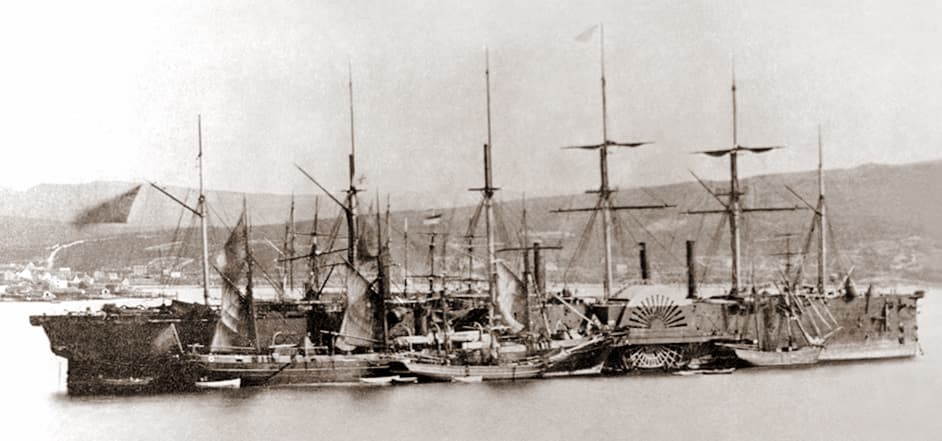Black and white photo of large ship with many masts.  Four smaller ships sit alongside the larger ship. There are buildings and hills pictured in the distance