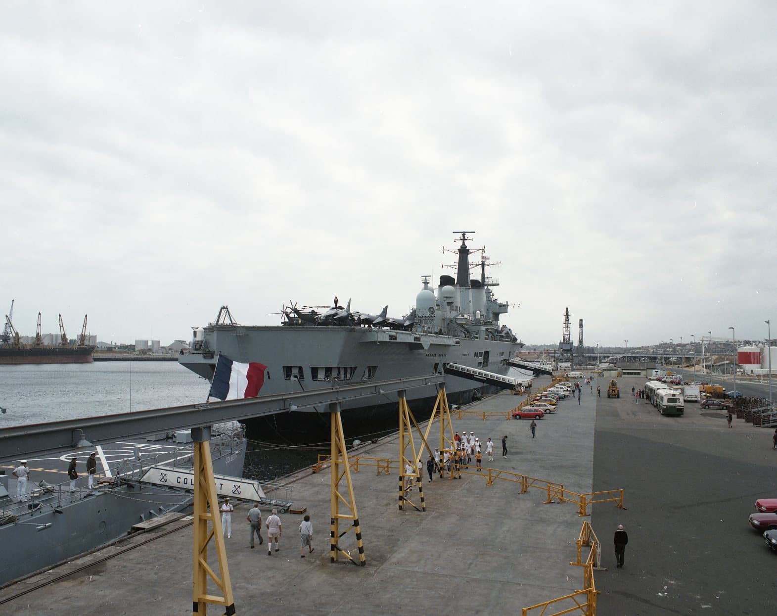 Colour photograph of a large aircraft carrier moored at a busy port under an overcast sky. The ship’s angled flight deck extends toward the pier, and aircraft and equipment are visible along the deck. A gangway connects the carrier to the wharf, where groups of people in uniform and casual clothing walk along the waterfront. Cranes, warehouses, and docked vessels fill the industrial harbour in the background.