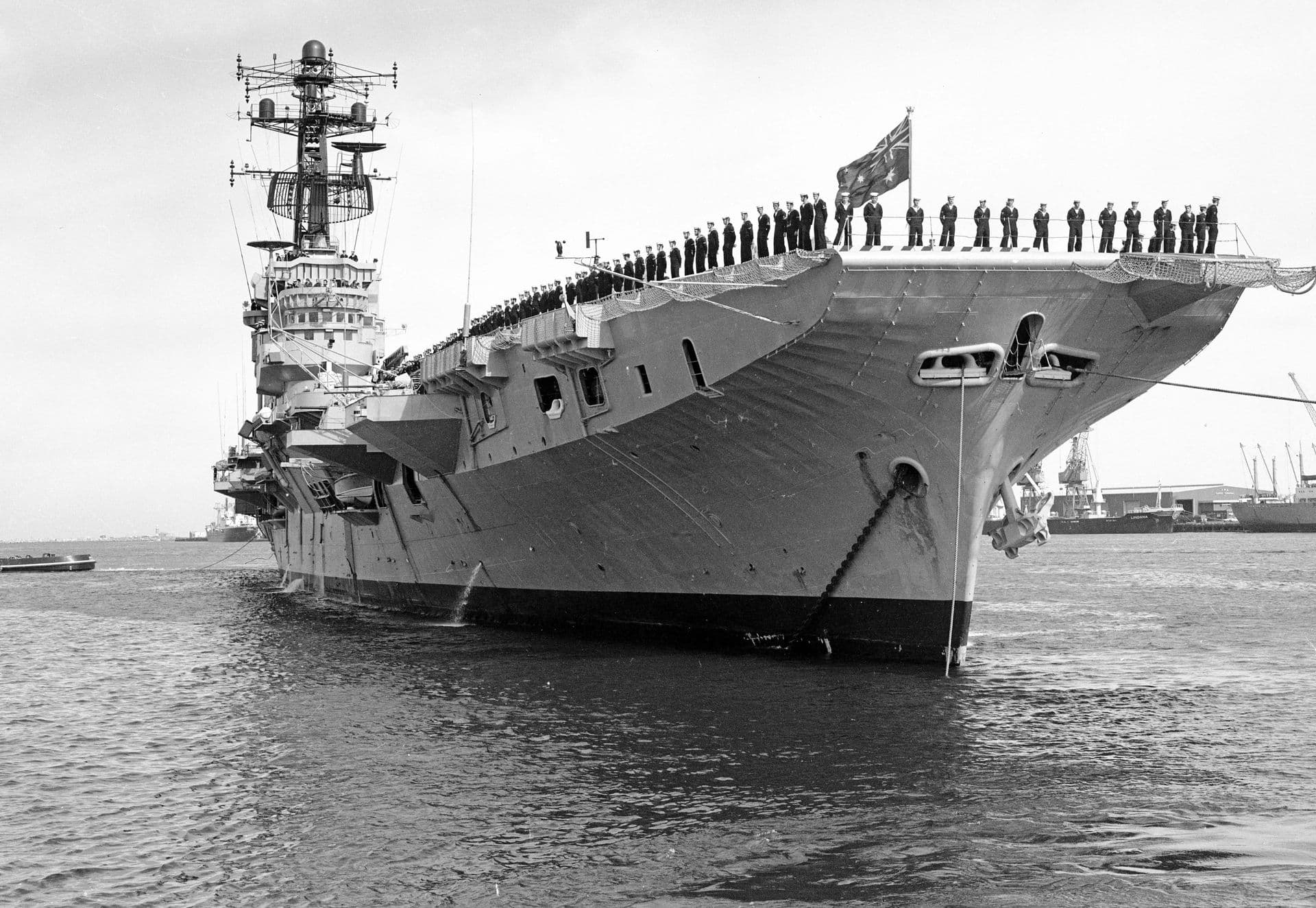 A large naval aircraft carrier is docked at a harbor, facing toward the viewer. Along the edge of its wide flight deck, rows of uniformed sailors stand in formation, suggesting a ceremonial moment. A flag flies at the bow. The ship’s tall superstructure rises above the deck, covered with radar dishes and communication equipment. Industrial port buildings and other vessels appear in the background