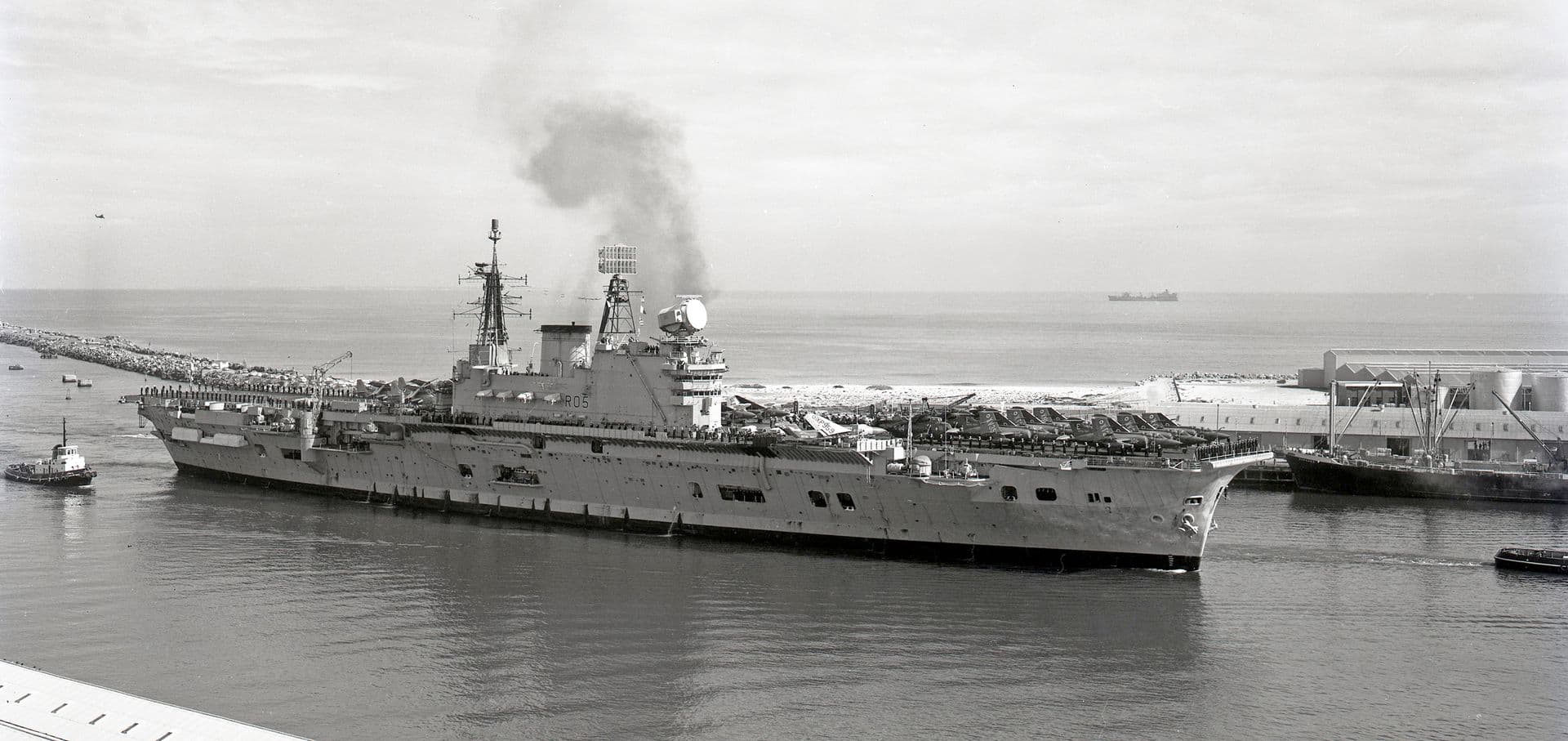 Black‑and‑white photograph of a large aircraft carrier entering a harbour, escorted by several tugboats. The ship’s flight deck is crowded with aircraft and equipment, and smoke rises from its funnel. People line the deck along the port side, watching the approach toward the breakwater and port facilities. The sea is calm, with another vessel visible on the horizon under a lightly overcast sky.