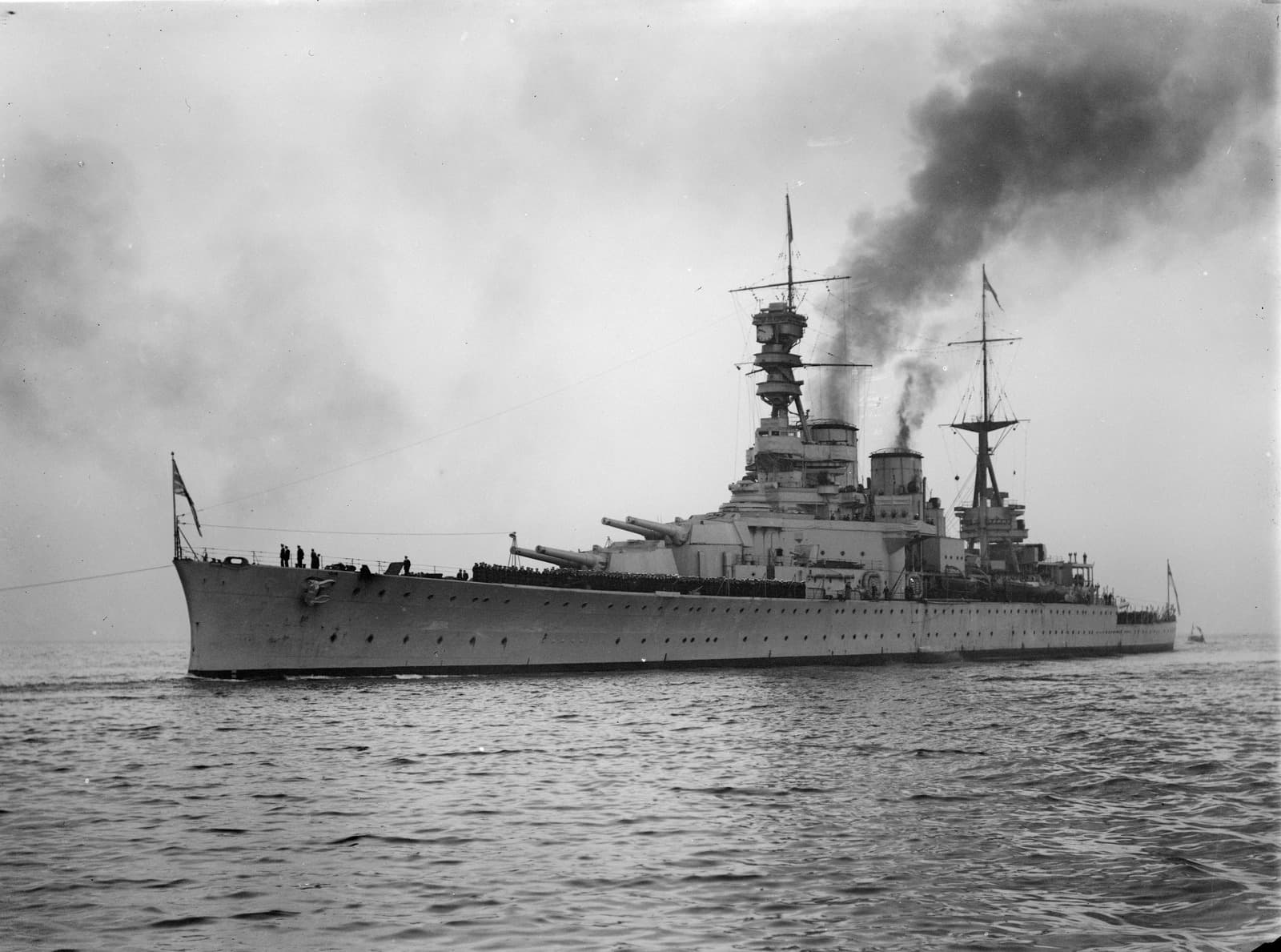 Black‑and‑white photograph of a large battlecruiser moving slowly across calm water. The ship’s long hull is lined with portholes and dominated by multiple large gun turrets. A tall, intricate superstructure rises above the forward deck, with masts, rigging, and lookout platforms visible. Two funnels toward the centre emit a dark plume of smoke that drifts across the sky. Crew members stand along the decks, and the distant horizon is hazy under an overcast sky.