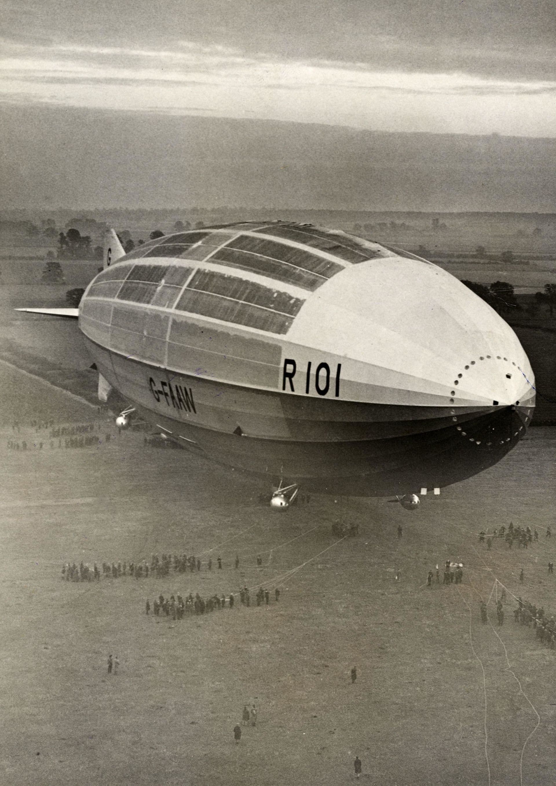 A black‑and‑white photograph of the large British airship R101 flying low over an open field. The airship’s streamlined hull dominates the frame, displaying its registration markings “R101” and “G‑FAAW” along the side. Several small ground handling crews and groups of spectators are visible far below, scattered across the field. The landscape stretches into the distance with trees, farmland, and a hazy sky at dawn or dusk.