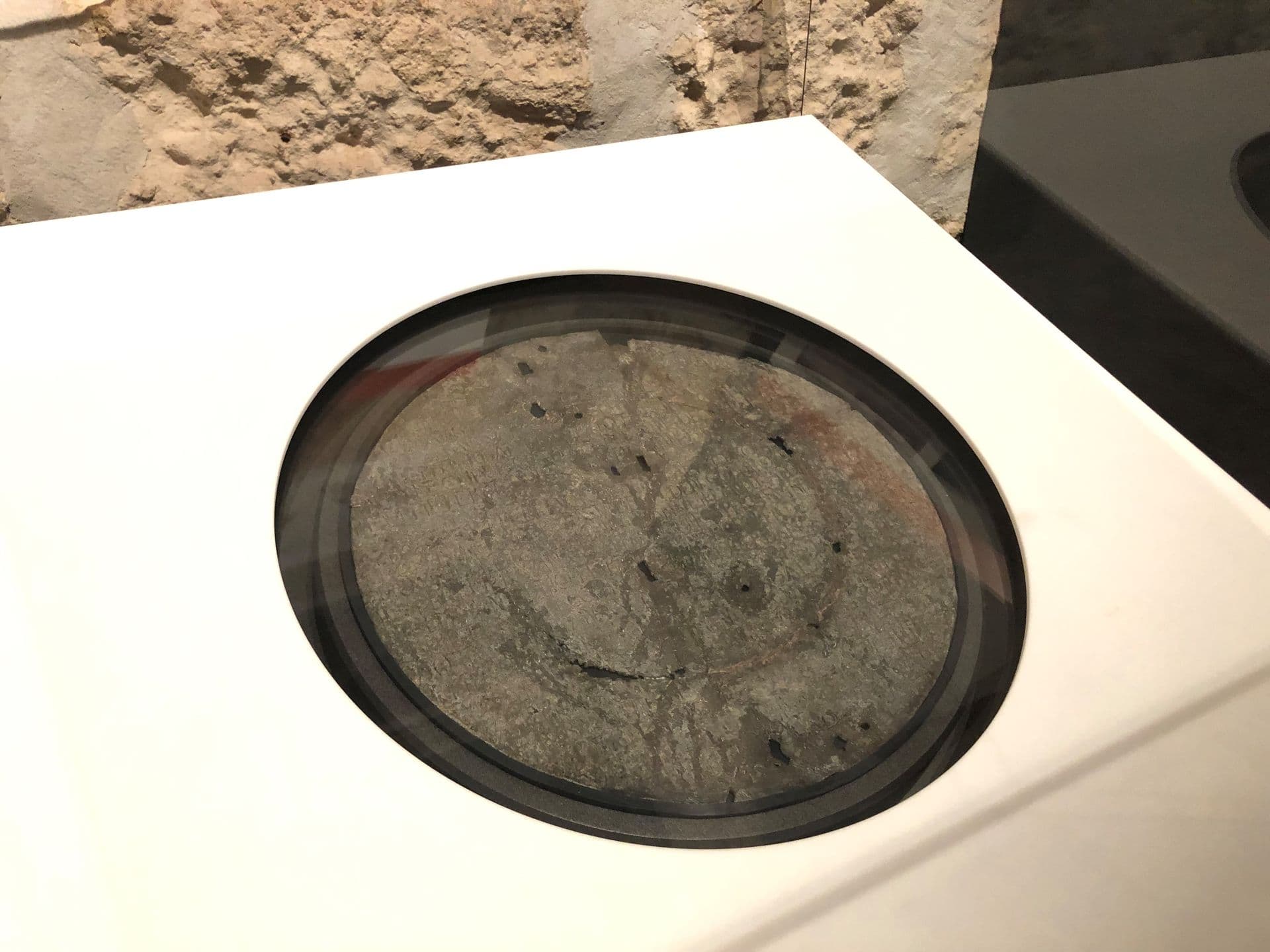 Round, grey, metal disc etched and engraved with lettering, and cracks and holes worn through age.