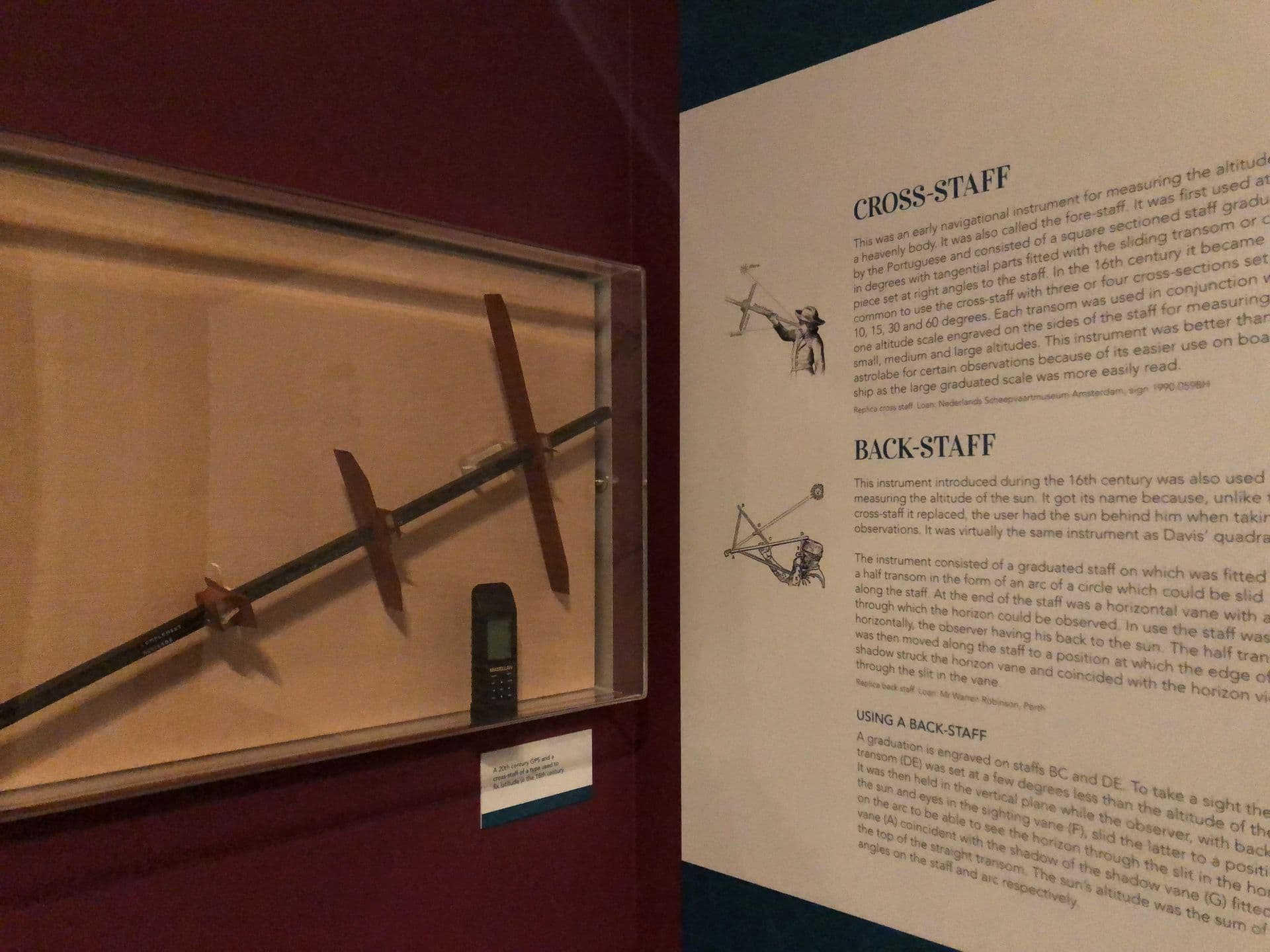 Long rod with two smaller pieces laid across mounted in a cabinet next to illustrations and information about Back Staff.