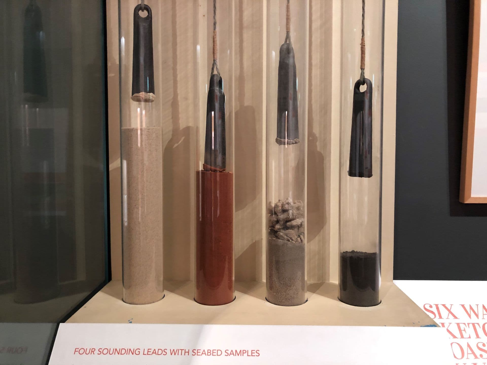 Cabinet containing four lead pieces suspended on string into glass containers with different samples of sea bed soils.
