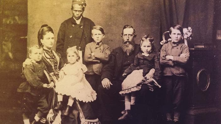 Old photo of family of 5 children and two adults seated, posing for portrait photo, and with one other gentleman standing behind them.
