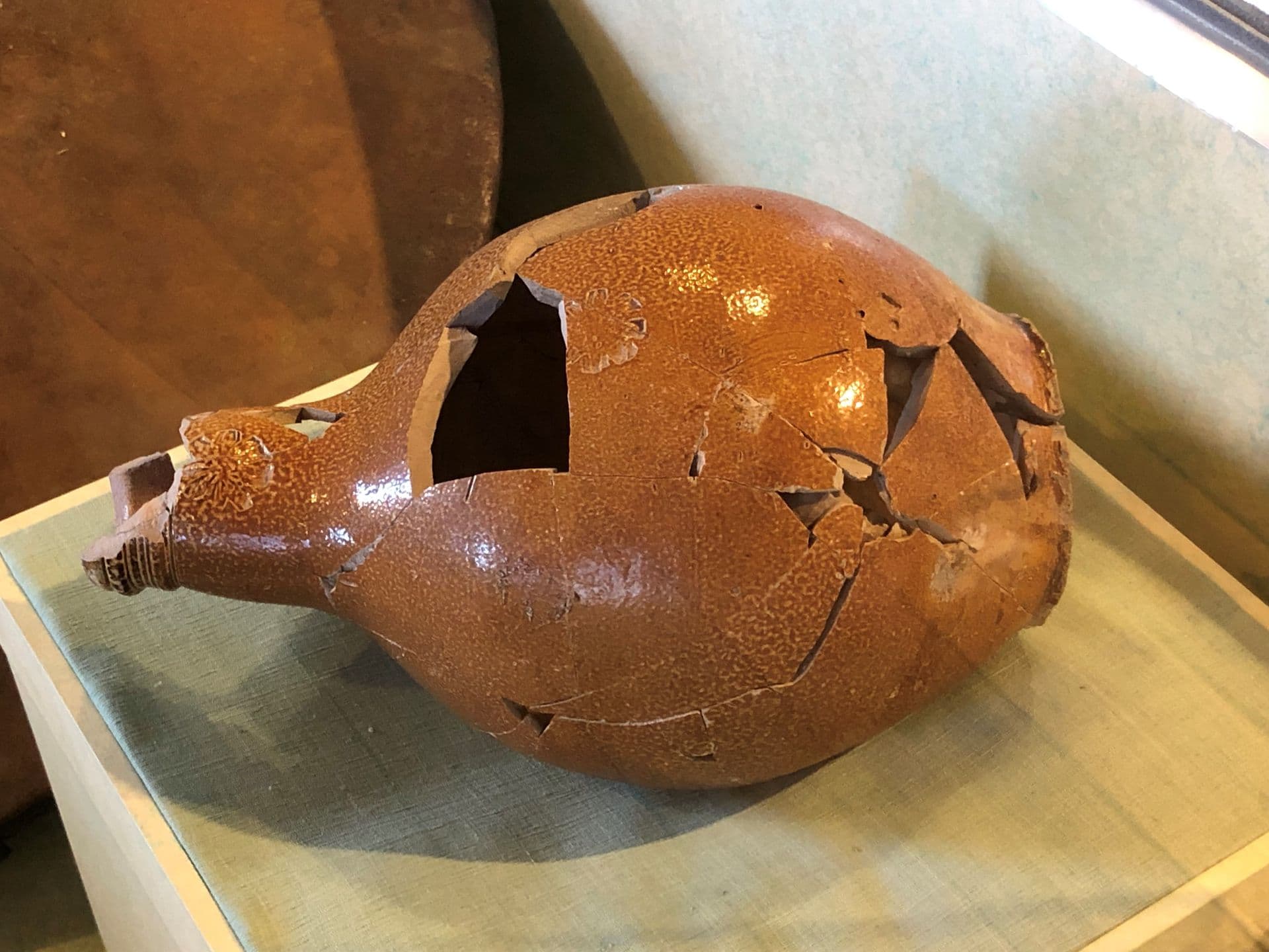 Orange and brown earthen wear jug, lying on it's side, that has been broken, and put back together, and still with missing pieces.