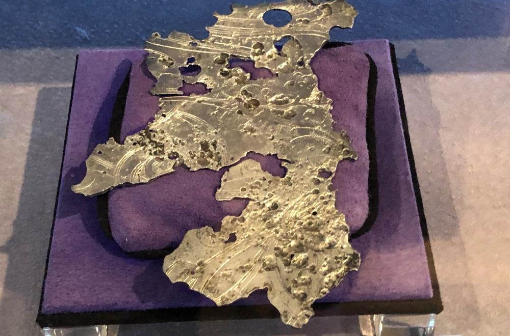 Jagged piece of silver metal with holes in it placed on a purple cushion in a display case.