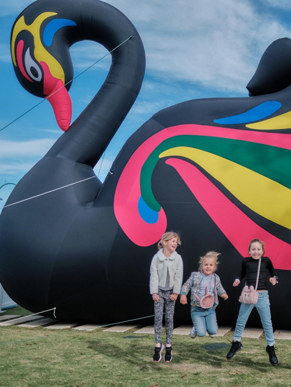Three kids jumping and laughing in front of an inflatable black swan in Fremantle 