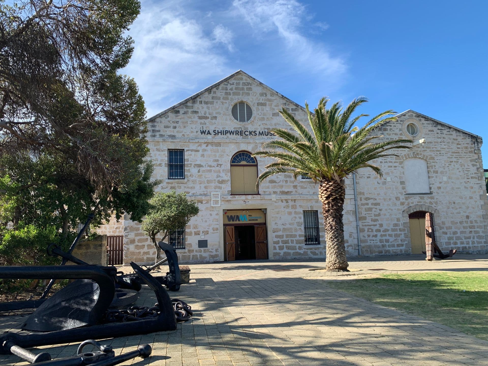 A building made of limestone bricks sits in afternoon shadow. Its forecourt is paved, and there is a palm tree in front of its wooden entry doors. Ships' anchors are on display to the left of the image.