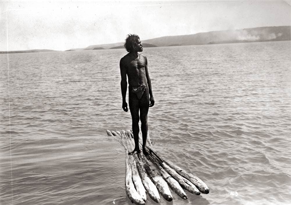 An aboriginal man stands tall on a raft on open water.