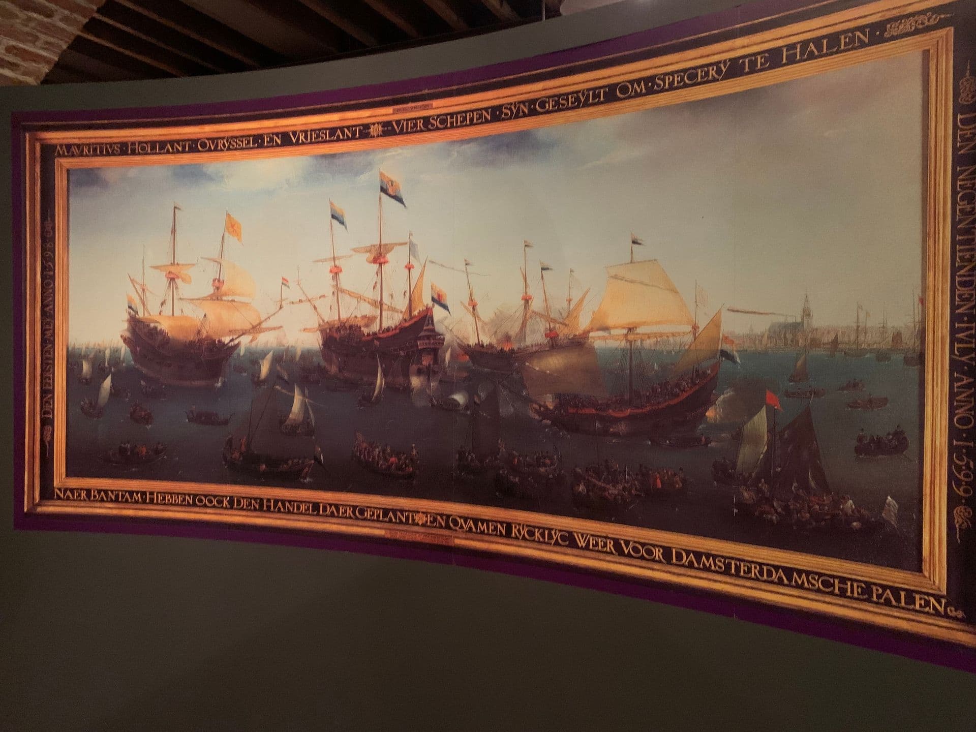 Four sailing ships with sails out and flags flying in port, surrounded by many smaller boats filled with people. Cannons have been fired, and in the distance can be seen a town with a church spire or town hall visible.