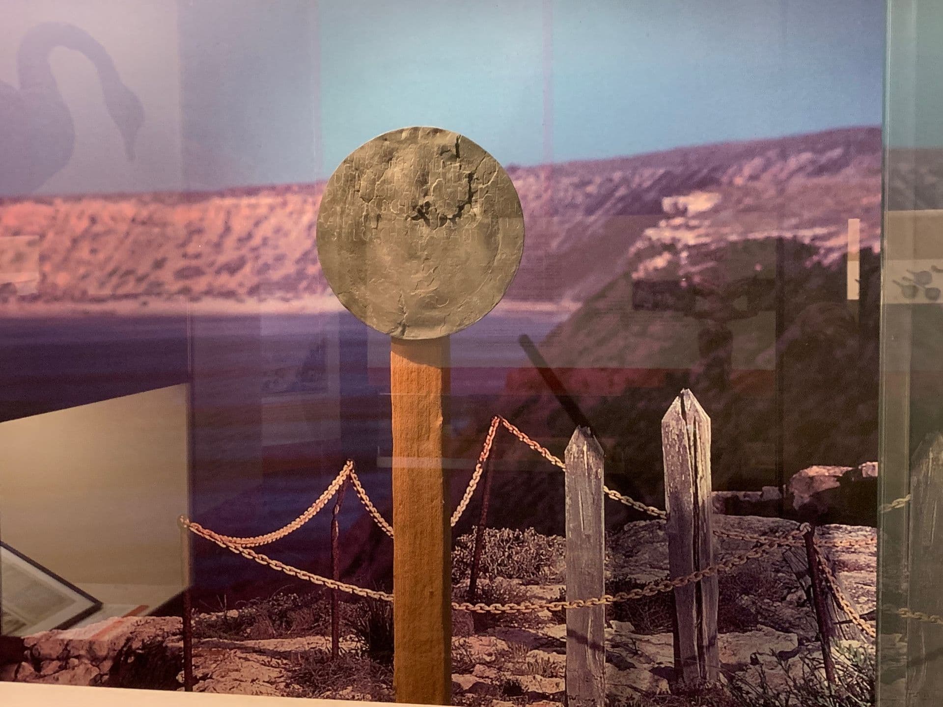 A replica of a flattened and engraved pewter dish, nailed to a wooden post, with a photographic backdrop of a rocky cliff at the edge of the ocean.