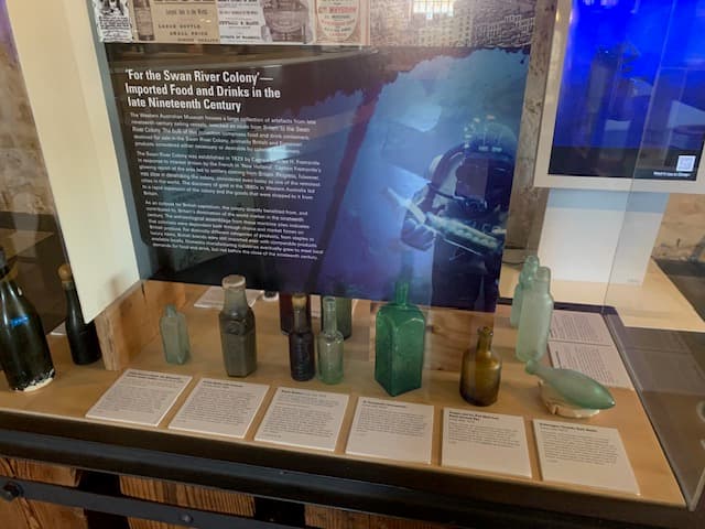 A photo of a display case featuring an information panel about imported food and drinks, and featuring glass bottles in assorted shades of clear, green and amber.