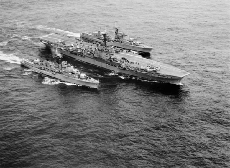 Three naval ships sailing together. The largest ship is in the centre and is carrying several aircraft.