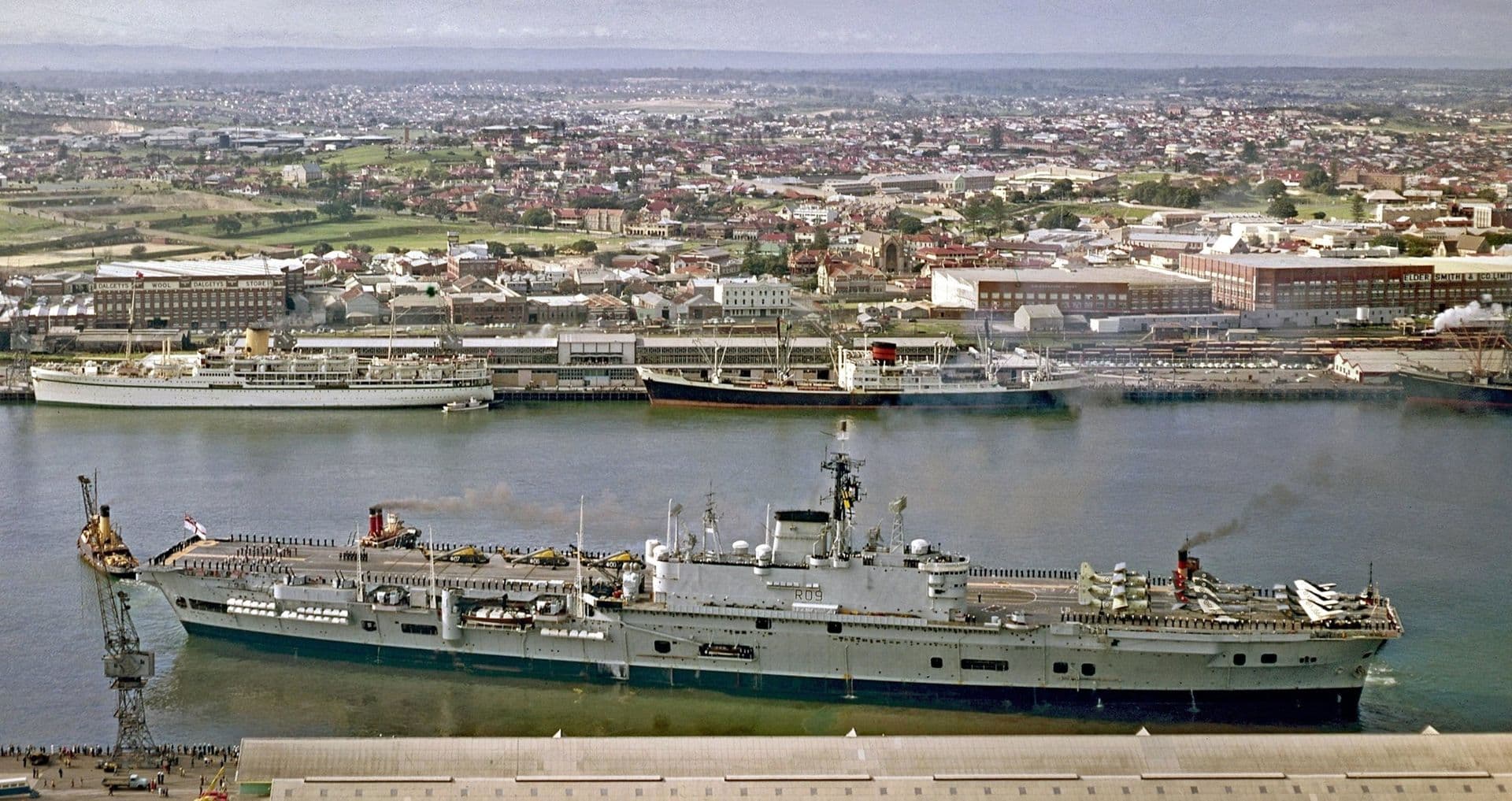 Colour aerial photograph of a large aircraft carrier moving through a harbour, escorted by tugboats. The ship’s deck is lined with personnel and equipment, and a trail of smoke rises from its funnels. Along the opposite shoreline are passenger ships docked beside a busy waterfront precinct, with warehouses, cranes, and industrial buildings visible. Beyond the port, a wide expanse of suburban houses stretches into the distance under a hazy sky.