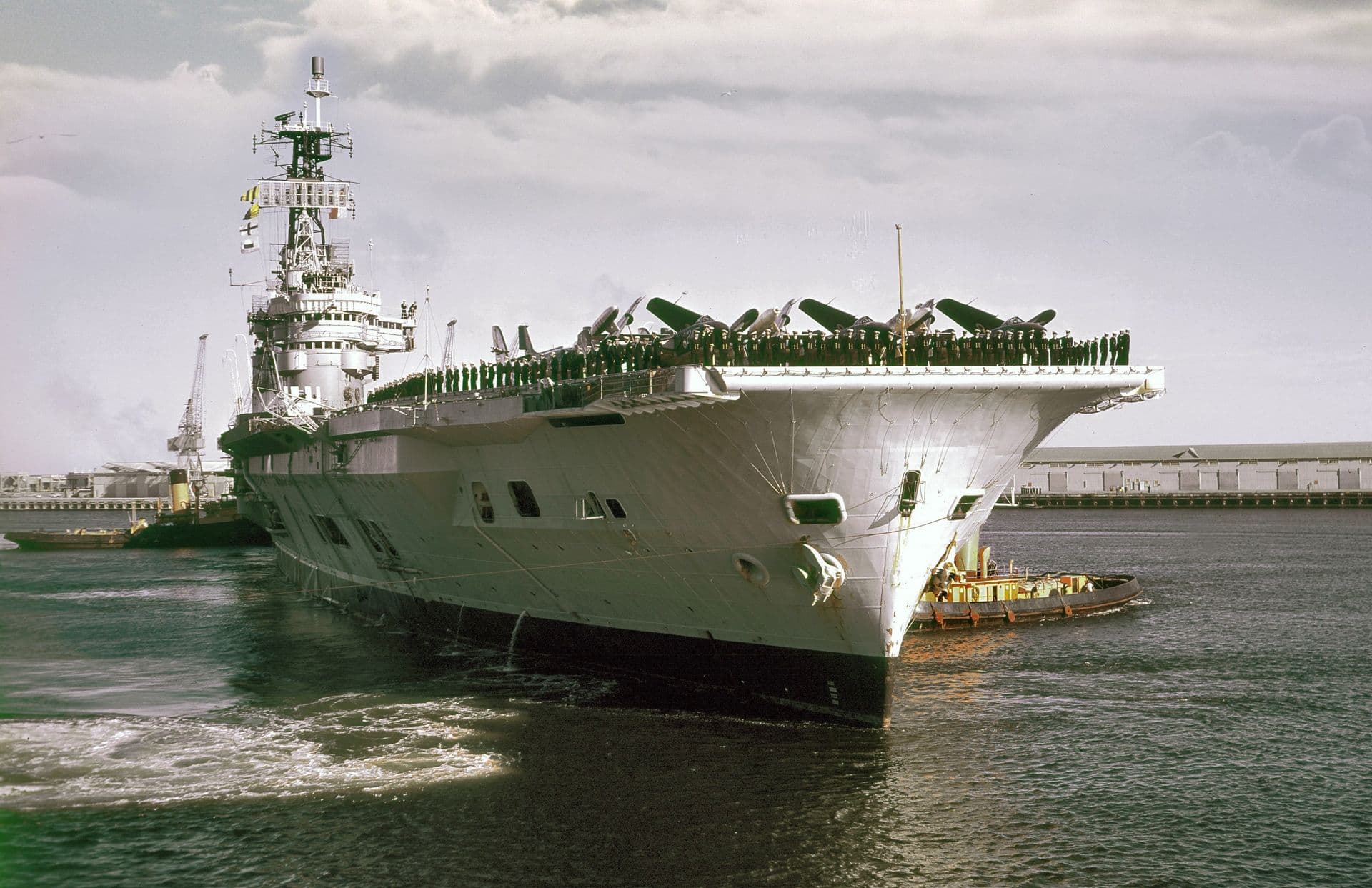 Colour photograph of a large aircraft carrier turning in a harbour, assisted by a tug positioned alongside its bow. The ship’s massive flight deck overhangs the water, lined with personnel standing along the edge. The superstructure rises high above the deck, fitted with radar masts and communication equipment. In the background are port buildings and cranes under a cloudy sky, while the water churns around the ship as it manoeuvres.