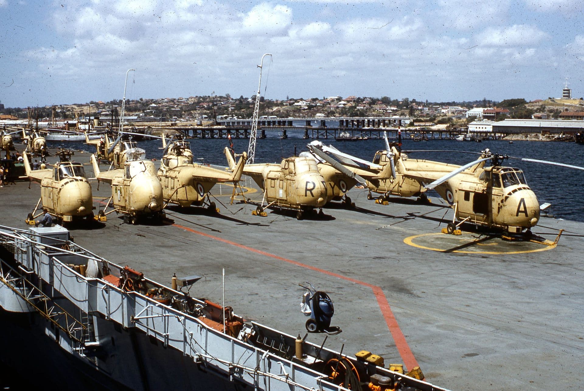 Colour photograph showing a row of light‑coloured naval helicopters parked on the flight deck of an aircraft carrier. The helicopters are arranged neatly with their rotors folded, and deck markings in red and yellow are visible beneath them. In the background, a harbour scene unfolds with calm blue water, a long jetty, and a shoreline of buildings and houses under a partly cloudy sky.