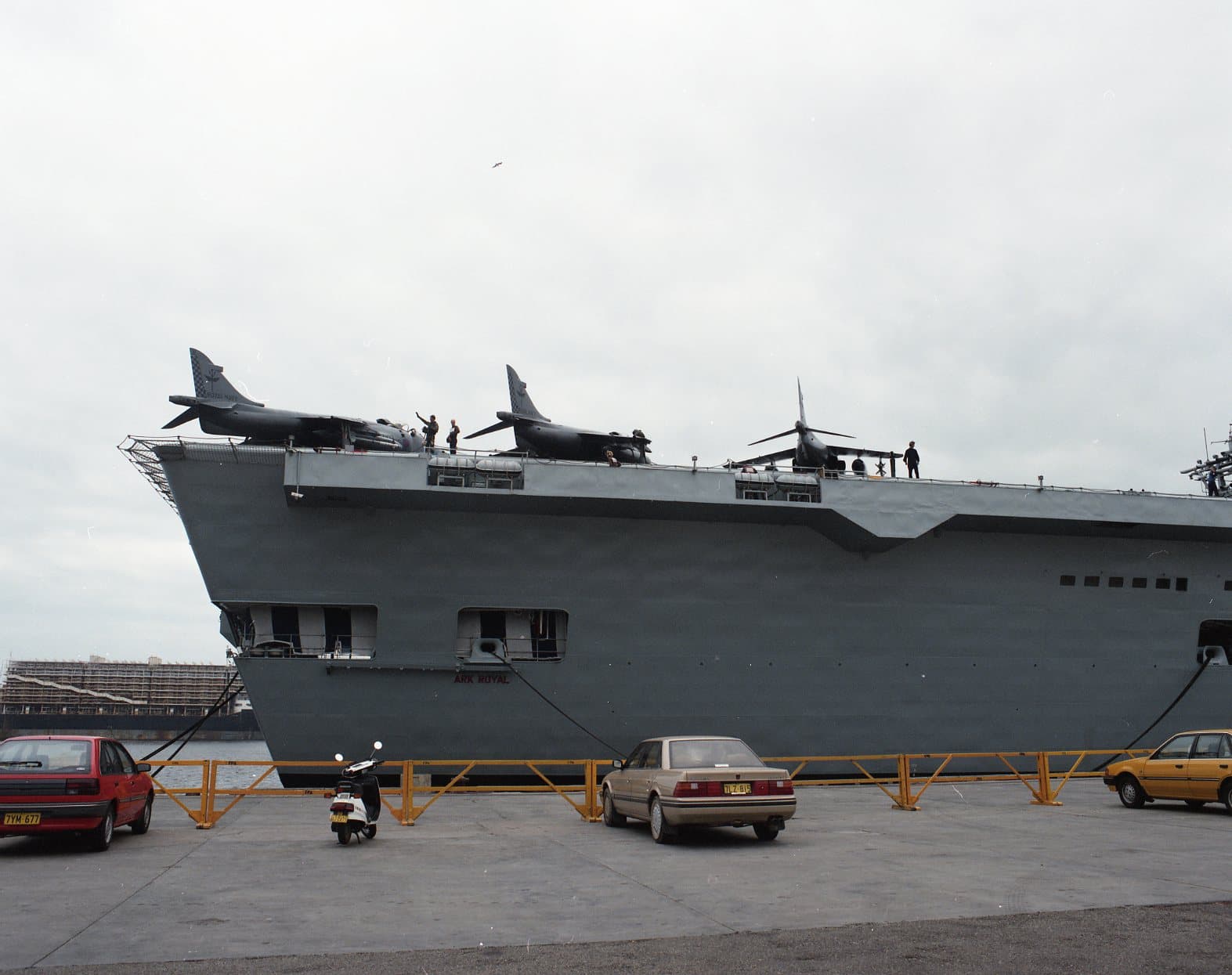 Colour photograph of the bow section of an aircraft carrier moored at a wharf. Several jet aircraft are parked along the forward edge of the flight deck, with crew members walking and working nearby. Below the overhang of the deck, the ship’s grey hull rises high above the waterline. Cars and a scooter are parked on the wharf in the foreground, and the sky is overcast.