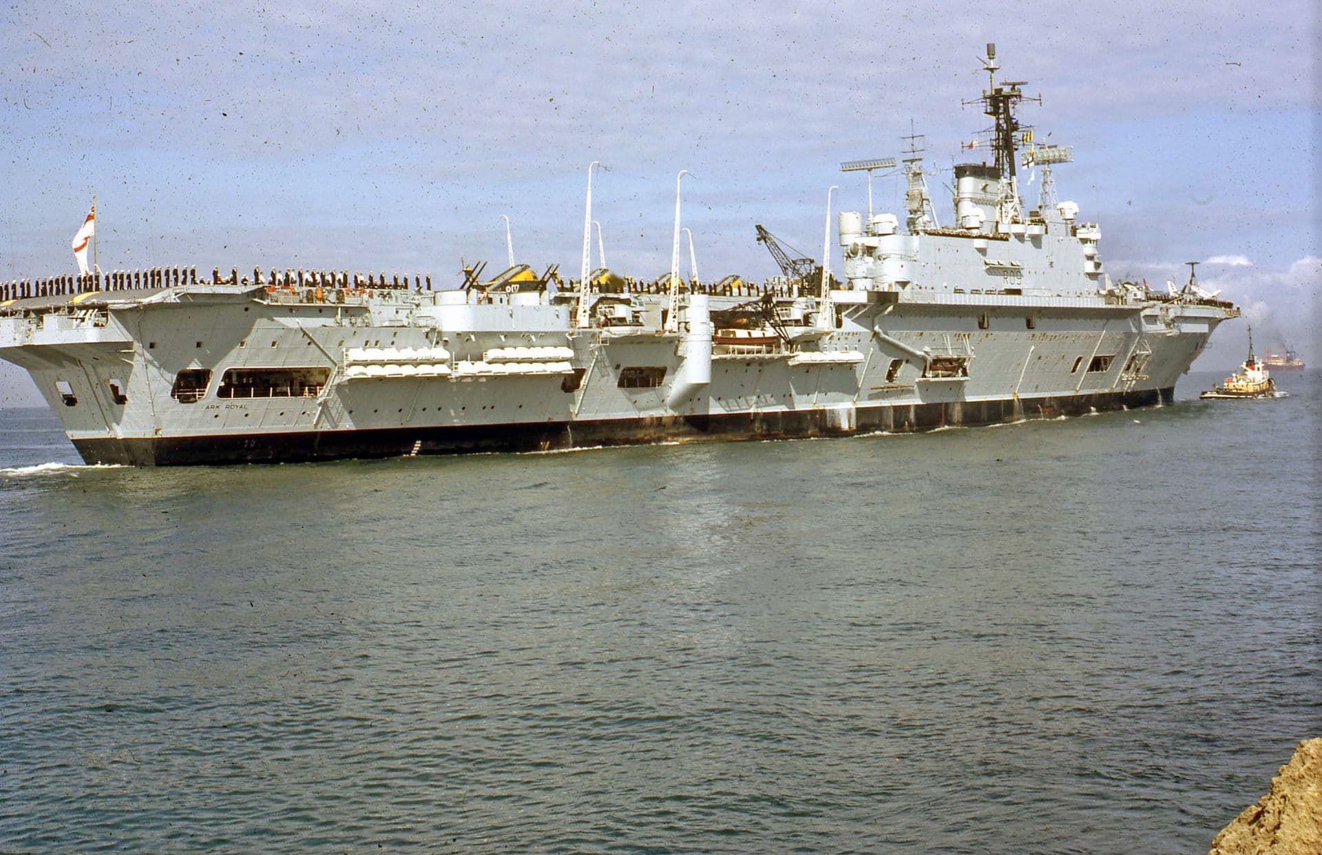 Colour photograph of a large aircraft carrier sailing out of a harbour. The ship’s deck is lined with personnel standing along the edge, and aircraft and equipment are positioned toward the rear of the vessel. The superstructure rises high above the flight deck, fitted with radar arrays and communication masts. A tugboat follows close behind on the right, and the ship moves through calm water under a partly cloudy sky.