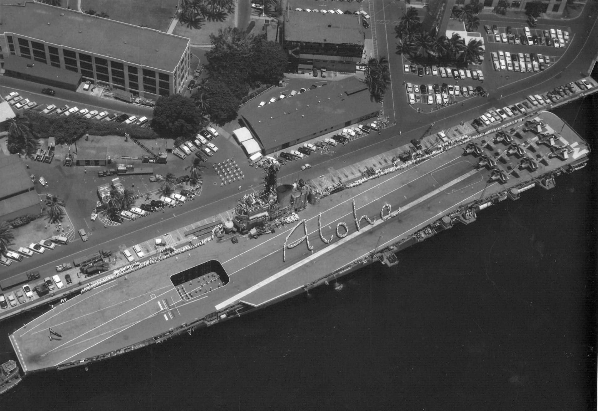 An overhead black‑and‑white photograph of a large aircraft carrier docked at a waterfront. The ship is moored beside a busy port area with cars, buildings, and palm trees visible along the shoreline. On the flight deck, crew members have arranged themselves to form the word “Aloha” in large cursive letters. The scene captures a welcoming or ceremonial moment as the ship rests in calm harbor waters.