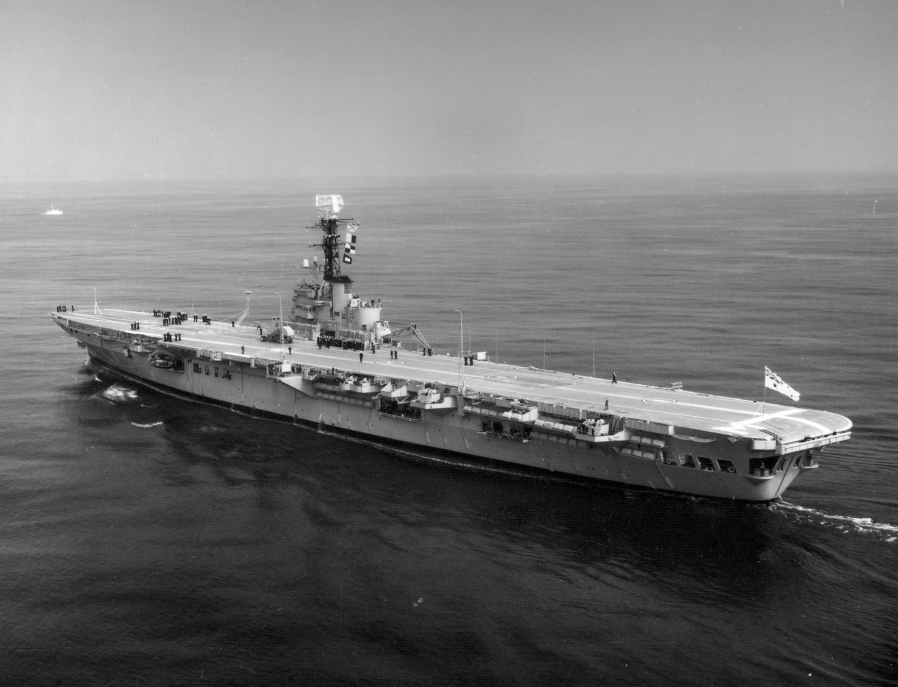 An aerial black‑and‑white photograph of a large aircraft carrier moving steadily across calm open water. The ship’s long, flat flight deck stretches almost the full length of the image, with several small groups of crew members visible along the deck. A tall island structure rises near the starboard side, topped with radar equipment and communication masts. Gentle wake trails behind the vessel as it travels through the sea under a clear sky.