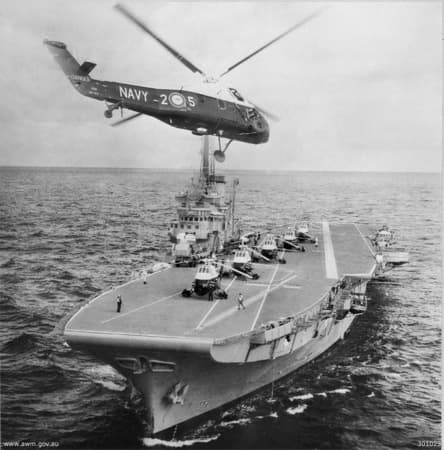 Black‑and‑white photograph of an aircraft carrier at sea with several helicopters parked on its flight deck. Another helicopter hovers just above the ship, aligned over the deck with its rotors spinning. Crew members stand near the parked aircraft, and the ocean stretches into the distance under a clear horizon.