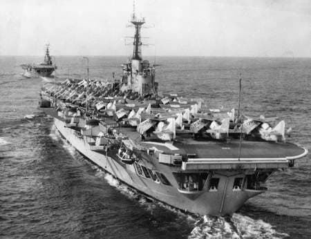 Black‑and‑white photograph showing an aircraft carrier at sea, viewed from behind and slightly above. The flight deck is packed with rows of parked aircraft facing forward. A second aircraft carrier follows in the distance along the same course. Both ships leave narrow wakes as they move across the open water under a clear sky.