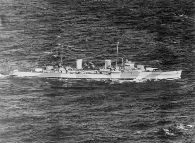 Black‑and‑white aerial photograph of a naval cruiser moving through choppy open water. The ship has two prominent funnels, multiple gun turrets positioned along the deck, and a patterned camouflage paint scheme along the hull. The vessel cuts through the waves, leaving a faint wake as it travels across the sea.