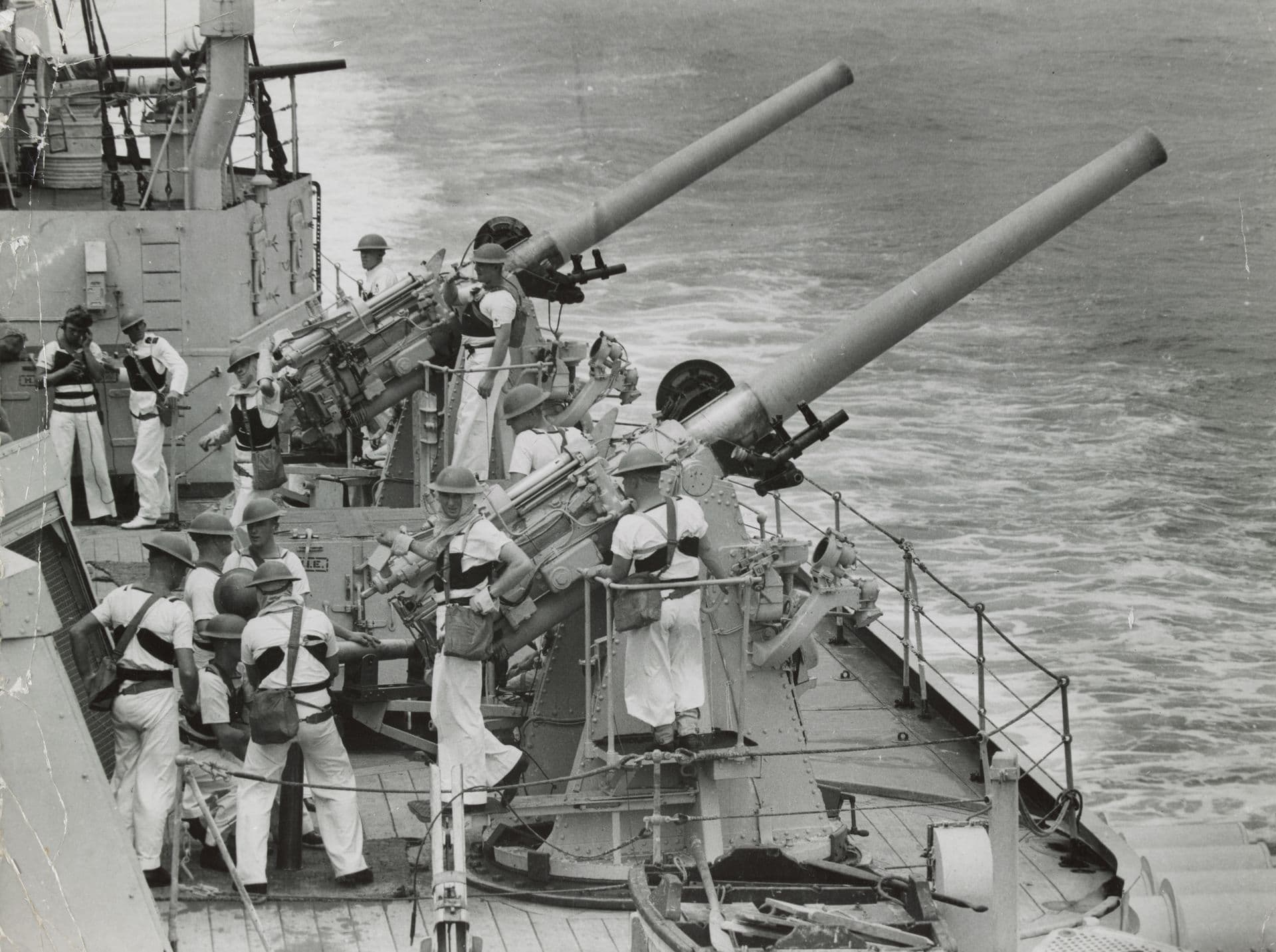 Black‑and‑white photograph showing a group of sailors operating anti‑aircraft guns on the deck of a naval ship. The crew, dressed in light uniforms and helmets, stand around several mounted guns pointing outward over the sea. Some sailors are adjusting equipment while others look toward the water. The ship’s wake is visible behind them as the vessel moves forward.