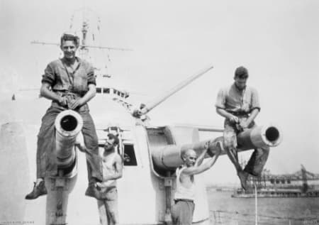 Black‑and‑white photograph showing sailors working on the barrels of large naval guns aboard a warship. Two sailors sit on top of the gun barrels while others stand beside or below them, cleaning and maintaining the equipment. The scene takes place on an open deck with parts of the ship’s superstructure and a distant shoreline visible in the background.
