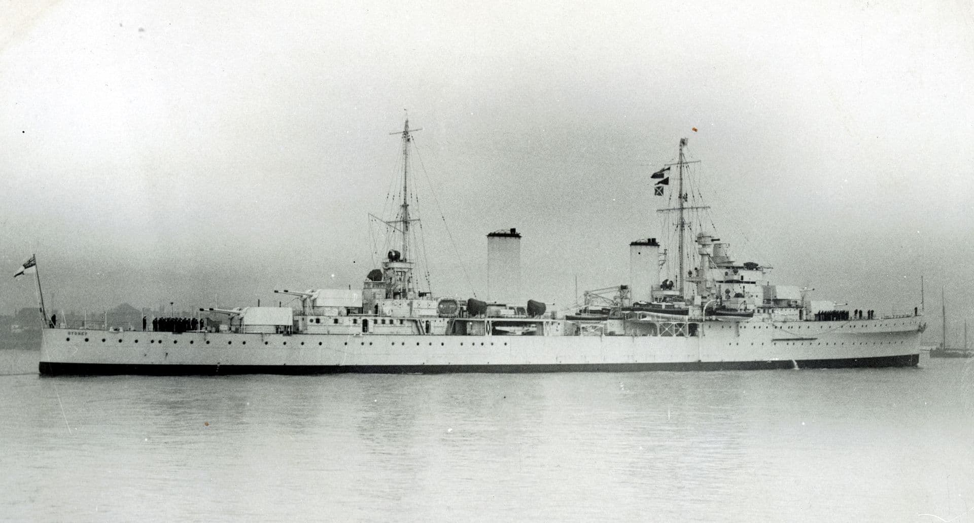 Black‑and‑white broadside photograph of a large naval cruiser at anchor or moving slowly across calm water. The ship has two tall funnels, multiple gun turrets along the deck, and prominent masts rising above the superstructure. A flag flies at the stern, and the background is hazy, with faint outlines of shoreline or other vessels barely visible through the mist.