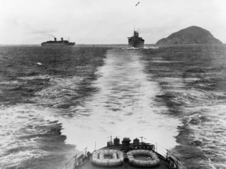 Black‑and‑white photograph taken from the stern of a ship looking out over the wake trailing behind it. Two large transport ships are visible ahead on the open sea, one to the left and one in the centre of the frame, both moving forward. A rocky island rises from the water on the right‑hand side in the distance. The scene shows choppy water, life rafts secured on the deck in the foreground, and a cloudy sky overhead.