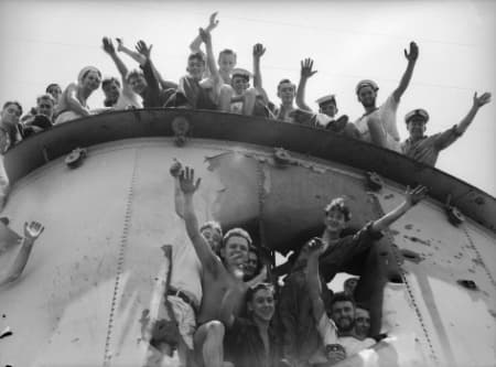 A group of sailors gather around and atop a large, battle‑damaged structure on a ship, smiling and waving toward the camera. Some stand at the rim while others lean out through openings in the metal, creating a lively, celebratory scene. The damaged surface of the structure is visible behind them, marked by dents and holes, while the sailors’ raised arms and relaxed poses convey a sense of camaraderie and relief.