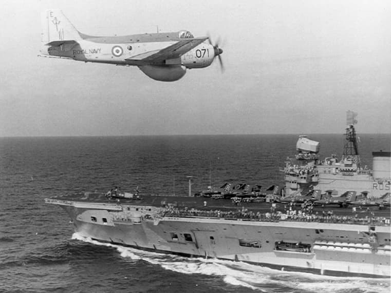 Black‑and‑white photograph of a propeller‑driven naval aircraft flying low over an aircraft carrier at sea. The aircraft, marked with Royal Navy roundels and the number “07,” is captured in level flight above the ship’s starboard side. Below, the carrier’s flight deck is lined with parked aircraft and personnel, with the island superstructure rising prominently above the deck. The surrounding ocean appears calm under an overcast sky.