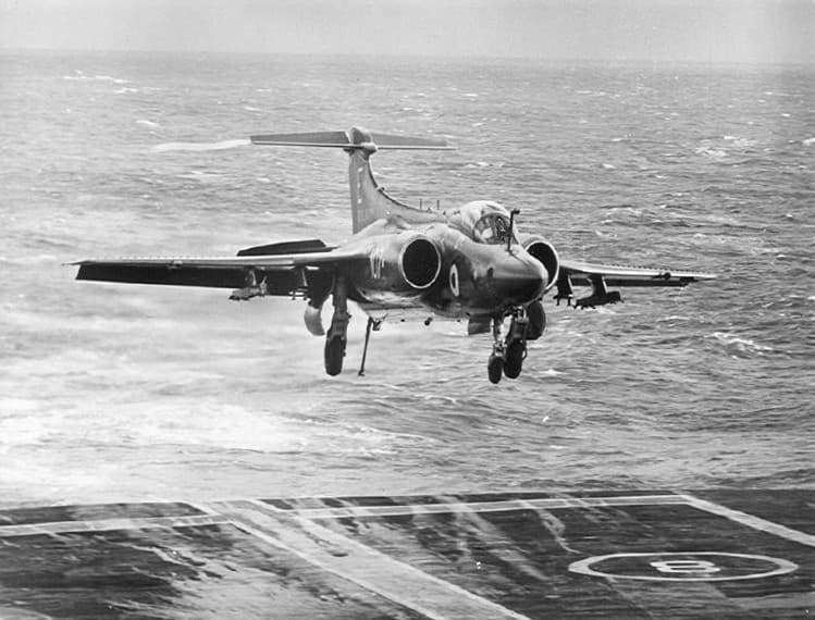 Black‑and‑white photograph of a naval strike aircraft approaching an aircraft carrier for landing. The aircraft, with its landing gear extended and wings spread, hovers just above the ship’s flight deck as waves churn below. The deck markings, including a large landing circle, are clearly visible in the foreground. The sea behind the aircraft appears rough, with whitecaps stretching toward the horizon under an overcast sky.