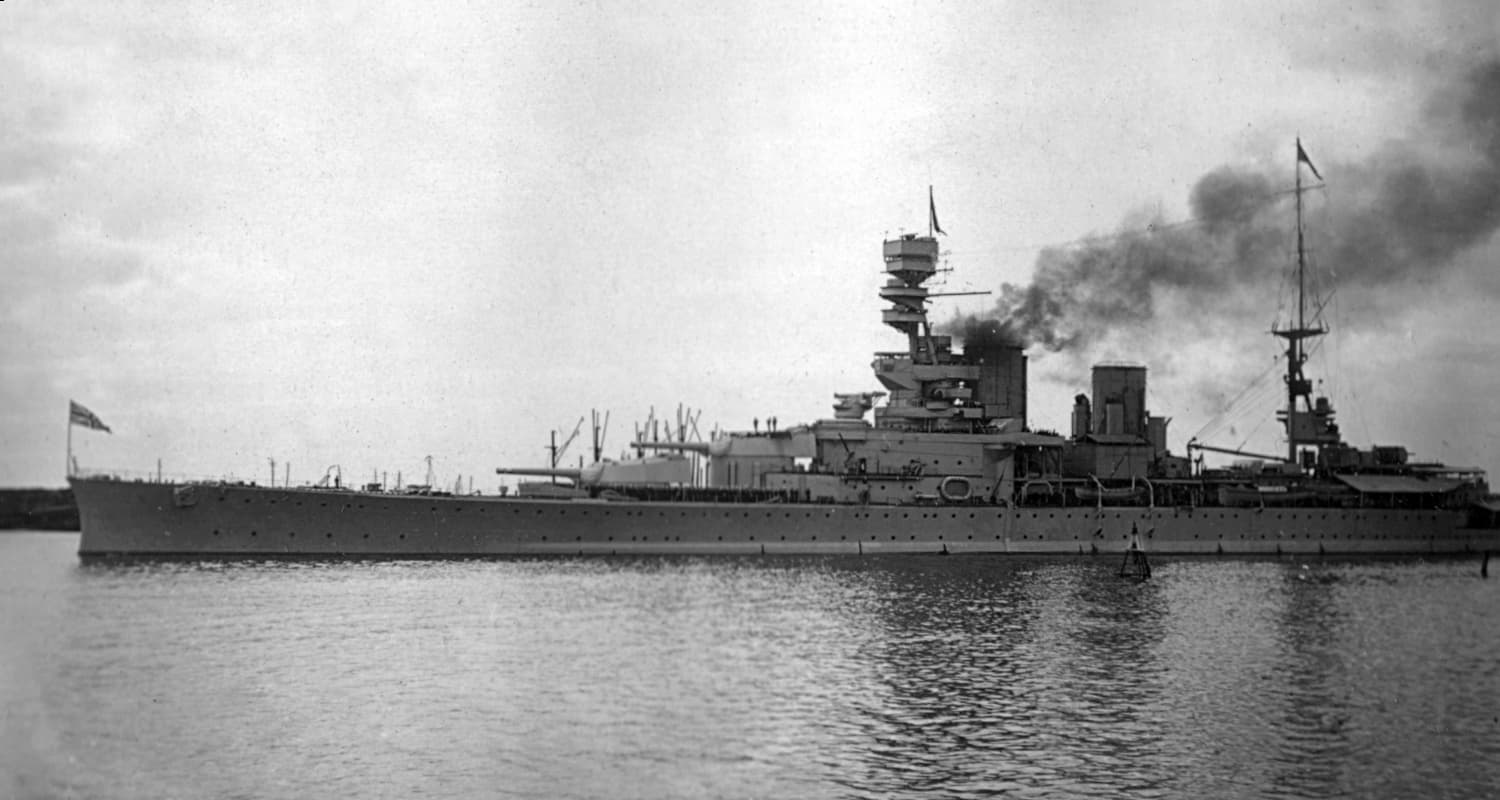 Black‑and‑white photograph of a large battlecruiser at anchor or moving slowly across calm water. The ship has a long, sleek hull and a tall forward superstructure topped with a distinctive fire‑control tower. Smoke rises from one of the funnels, drifting across the sky. Gun turrets are positioned along the deck, and a naval ensign flies at the stern. The background shows a low, hazy shoreline beneath an overcast sky.