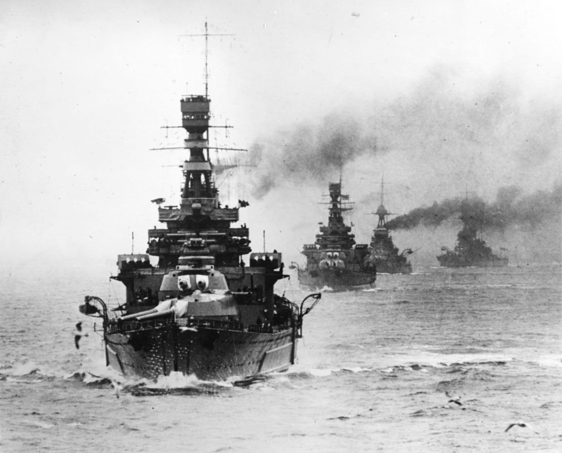 Black‑and‑white photograph showing a line of large battlecruisers or battleships underway at sea. The nearest ship is viewed from the front, cutting through the water with its bow creating a broad wake. Its tall superstructure, masts, and multiple gun turrets dominate the frame. Two more capital ships follow behind in formation, their funnels pouring dark smoke into the overcast sky. The sea is choppy, and the ships appear to be engaged in manoeuvres or a fleet exercise.