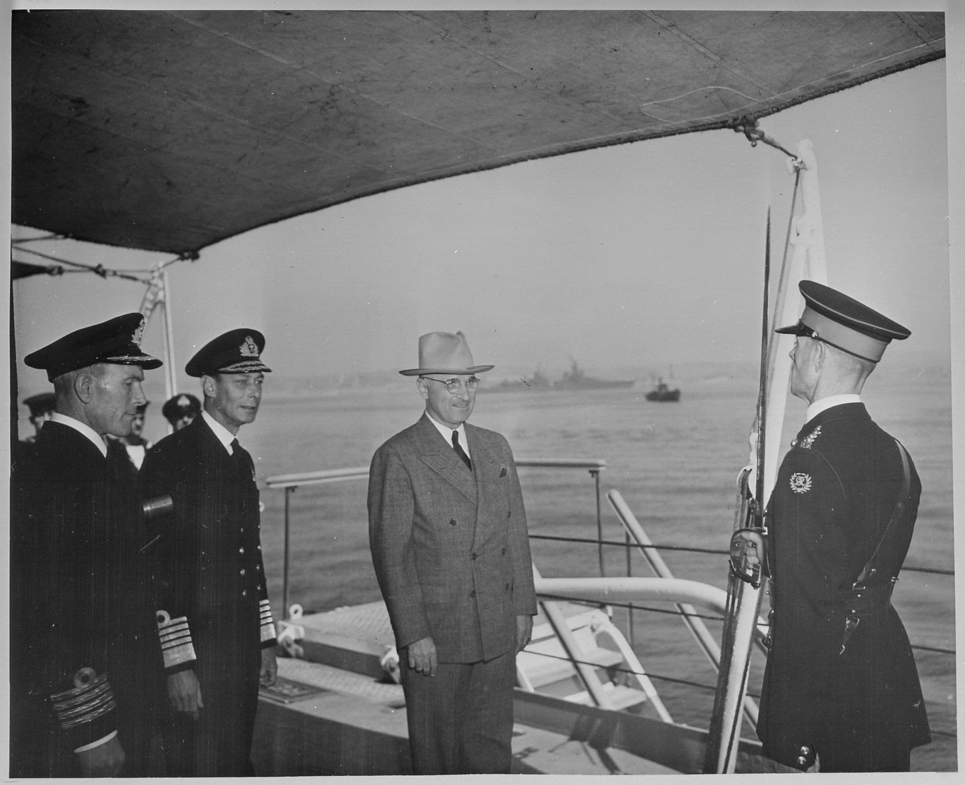 Black‑and‑white photograph showing a formal ceremony taking place on the deck of a naval ship. Several uniformed naval officers stand to one side, while a man in a suit and hat stands before an honour guard holding a rifle with a fixed bayonet. The group is positioned beneath a canvas awning. In the background, naval vessels and small craft are visible on the water, with a hazy shoreline beyond.