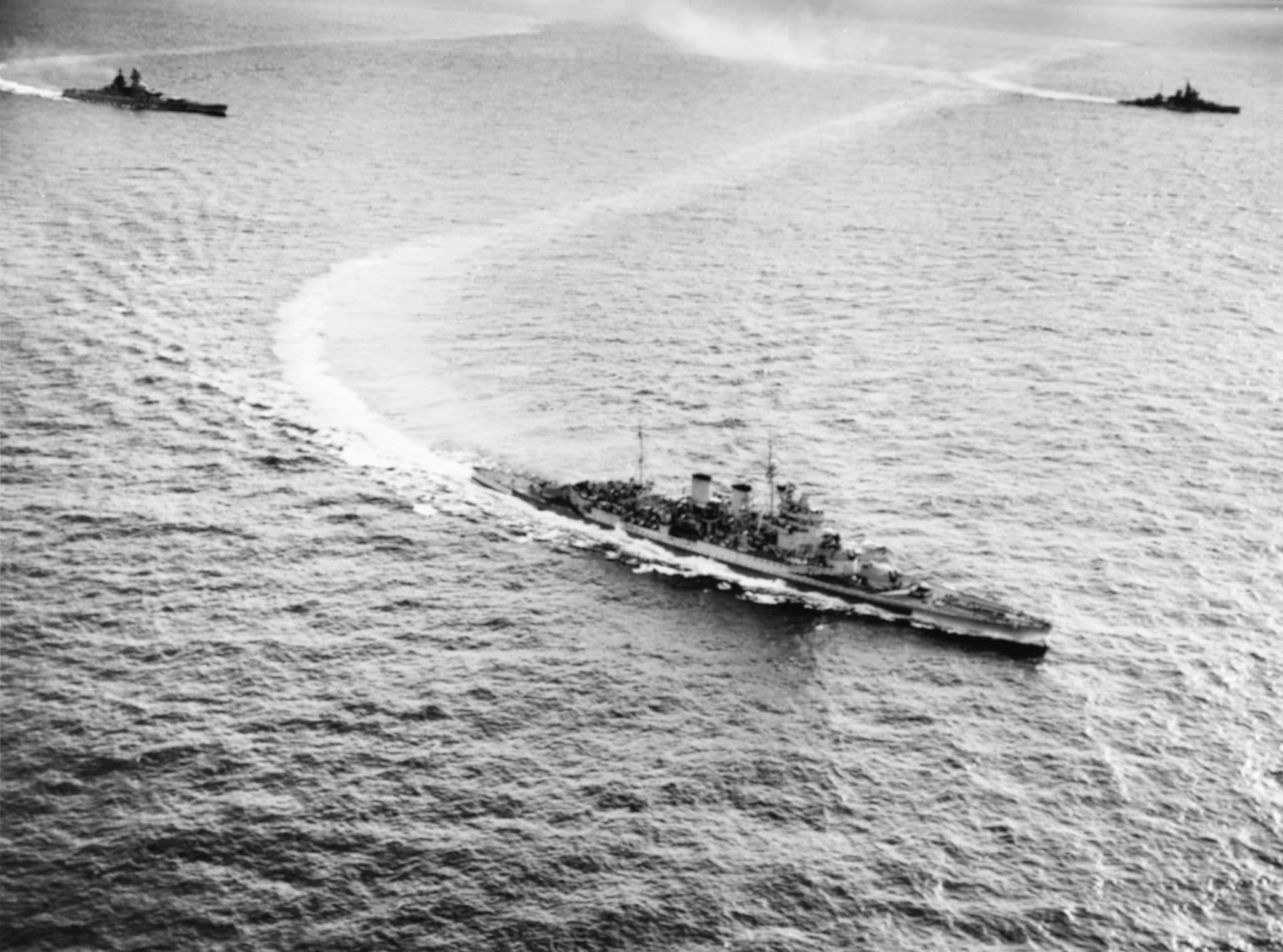 Black‑and‑white aerial photograph showing three large warships operating at sea in formation. The nearest vessel is a long, sleek capital ship turning sharply, leaving a sweeping white wake behind it. Two other battleships follow at wider distances, each creating faint wakes as they move across the open water. The surface of the sea is textured with small waves, and the ships appear to be conducting manoeuvres under an overcast sky.