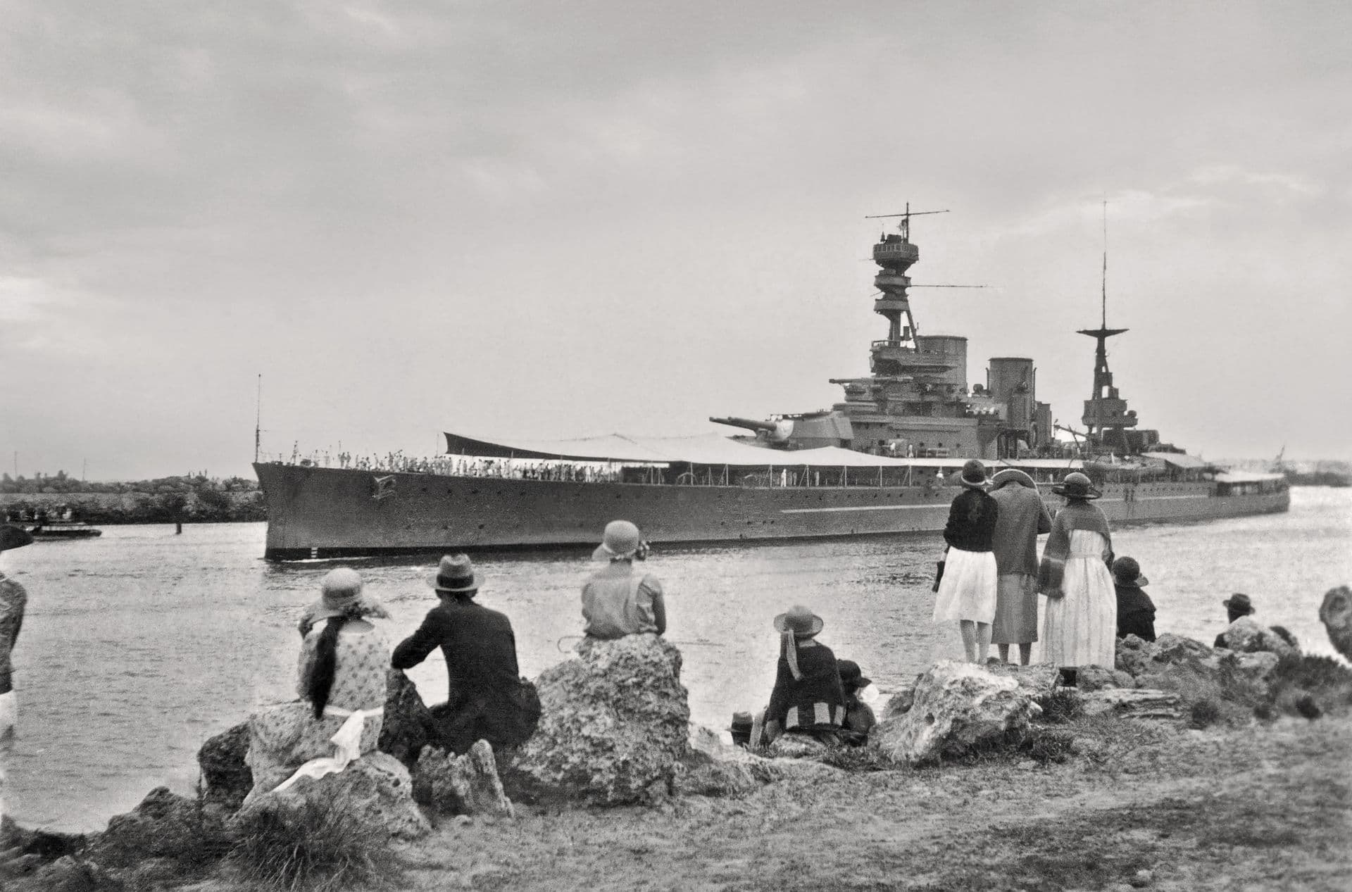 Black‑and‑white photograph showing a large battlecruiser moving slowly through a harbour channel. The ship’s long hull and tall superstructure dominate the scene, with awnings rigged along part of the deck and crew lined up at the rails. In the foreground, a group of people sit and stand on rocky ground near the water’s edge, watching the vessel pass. The landscape is flat and open, with low vegetation and a hazy sky overhead.