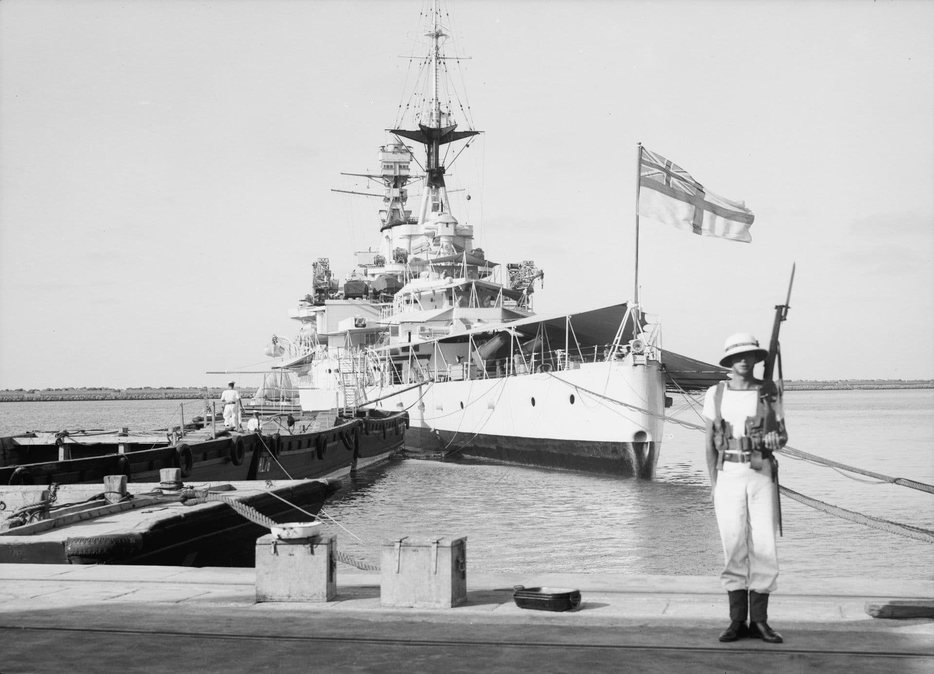 A black‑and‑white photograph of a large warship moored at a harbour pier. The ship’s bow faces the camera, showing its tall superstructure, multiple masts, and an ensign flying prominently from a flagstaff. Several small boats are tied alongside the vessel. In the foreground, a uniformed sentry stands on the quay holding a rifle, positioned near bollards and mooring lines. The water is calm, and the horizon stretches across a clear, lightly clouded sky.