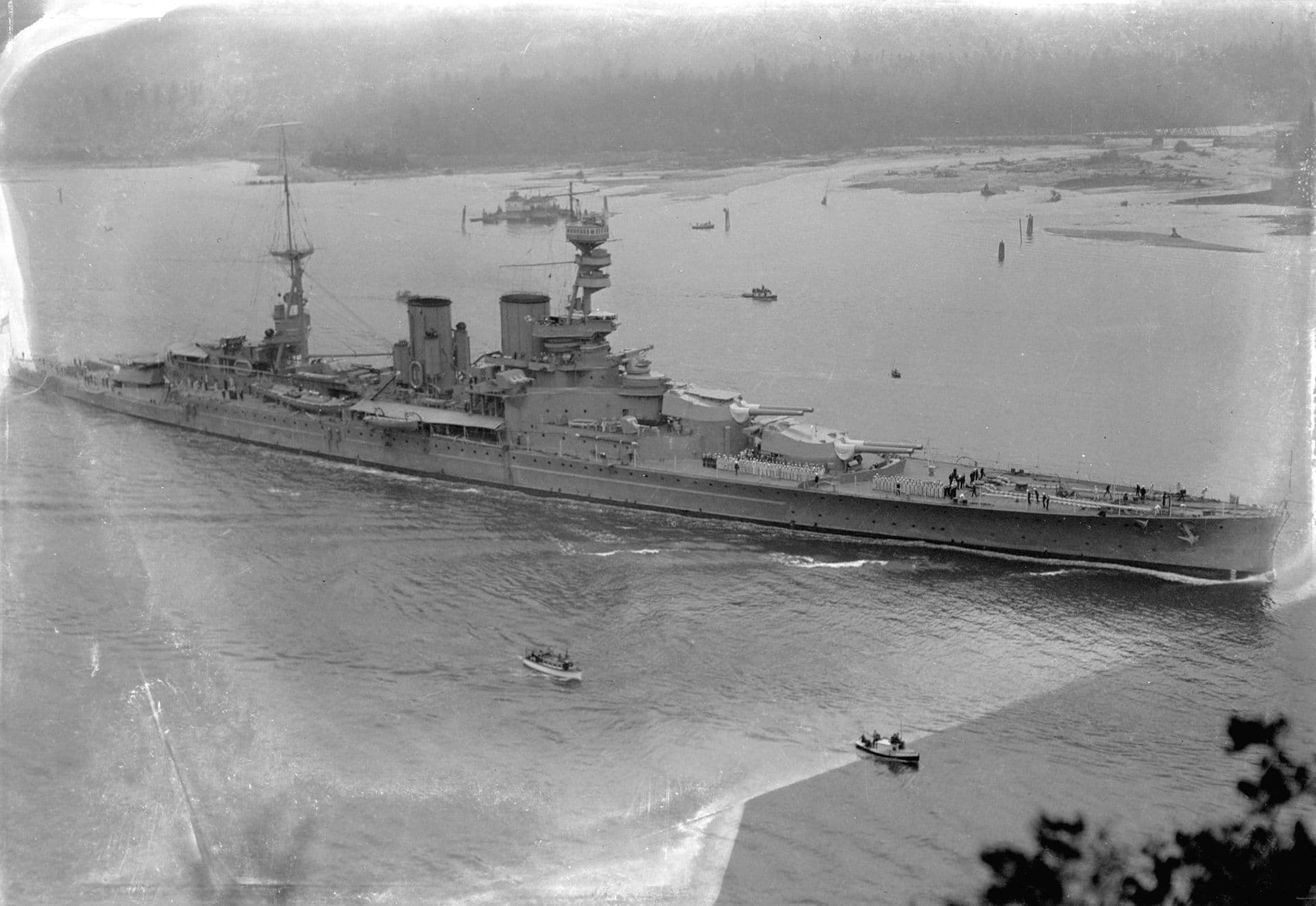 Black‑and‑white photograph of a large battlecruiser moving through a wide harbour channel, viewed from an elevated vantage point. The ship’s long hull, twin funnels, and tall forward superstructure are clearly visible, along with gun turrets positioned along the deck. Small motorboats travel nearby, leaving light wakes on the water. In the background, wooded shoreline and scattered vessels appear through a light haze.