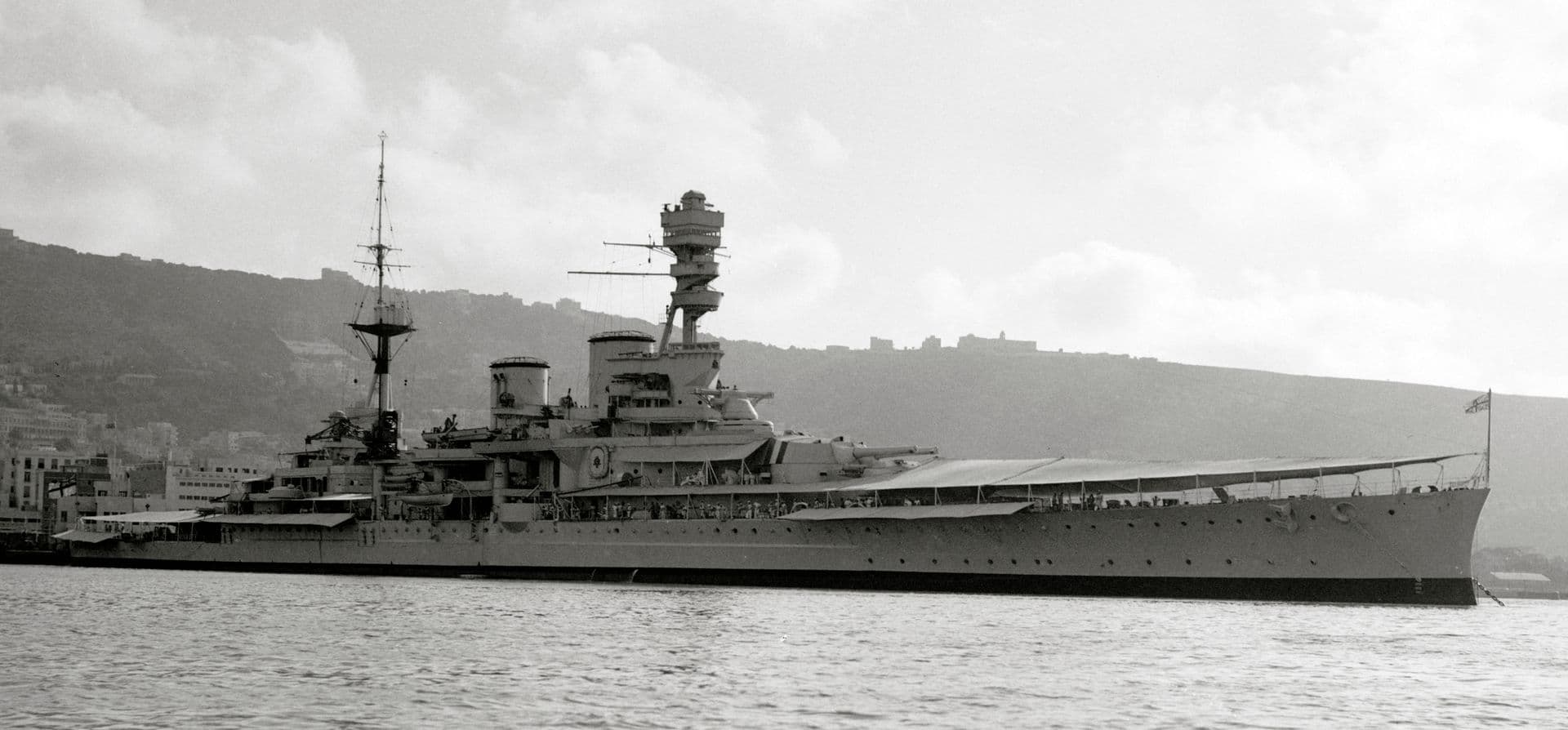 A black‑and‑white photograph of a large battlecruiser moored in a harbour, viewed broadside. The ship’s long, sleek hull features rows of portholes and several large gun turrets mounted forward. Its tall superstructure rises above the deck with masts, lookout platforms, and communication rigging clearly visible. A naval ensign flies at the stern. In the background, low hills and buildings line the shoreline beneath a cloudy sky.