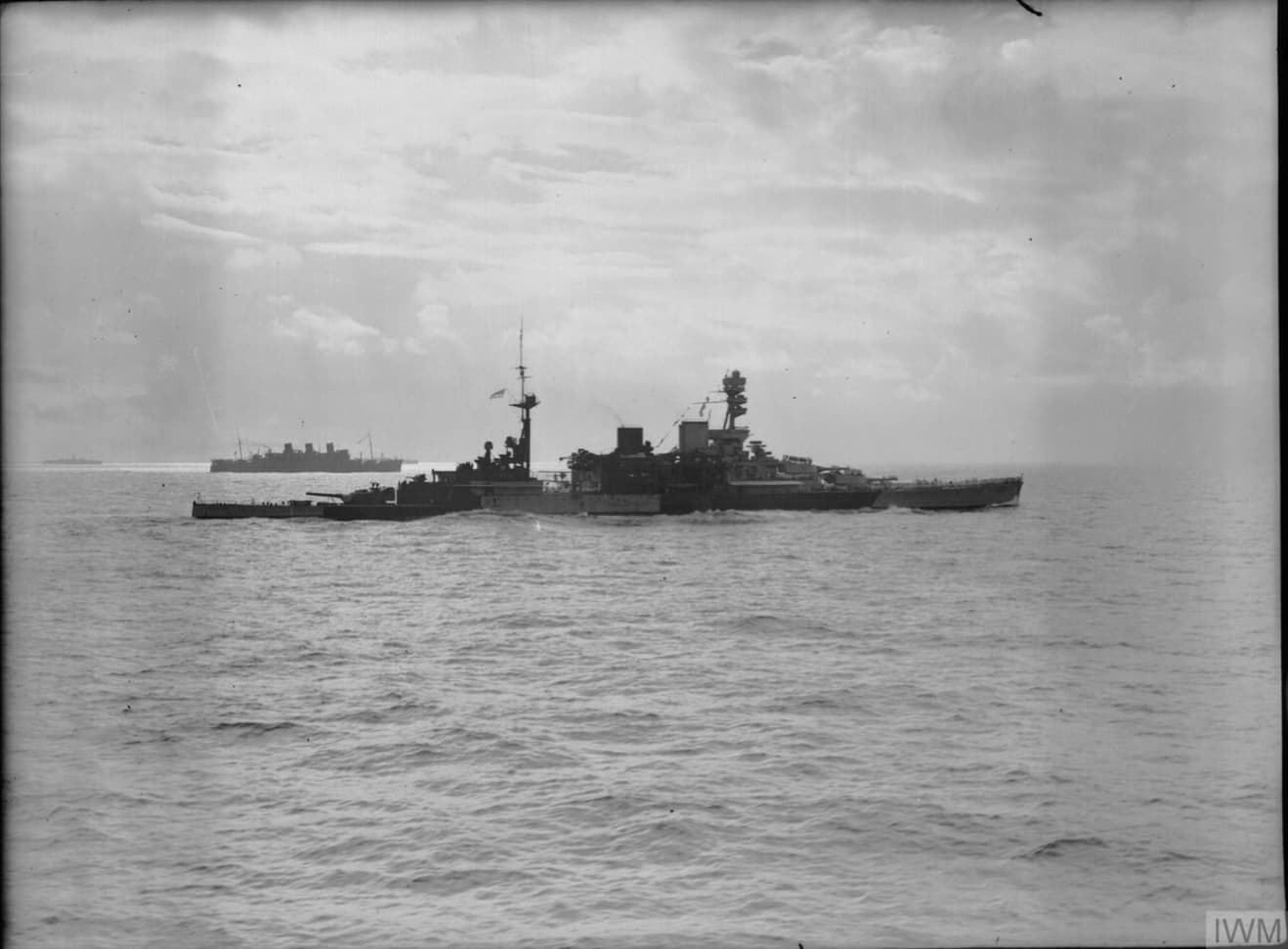 Black‑and‑white photograph of a battlecruiser at sea, viewed broadside from a distance. The ship’s long silhouette shows its superstructure, funnels, and gun turrets rising above the deck, while gentle waves surround the hull. On the horizon, several large troopships and other vessels travel in the same direction, partially obscured by haze. The sky is overcast with streaks of light breaking through the clouds, creating a subdued, open‑ocean scene.