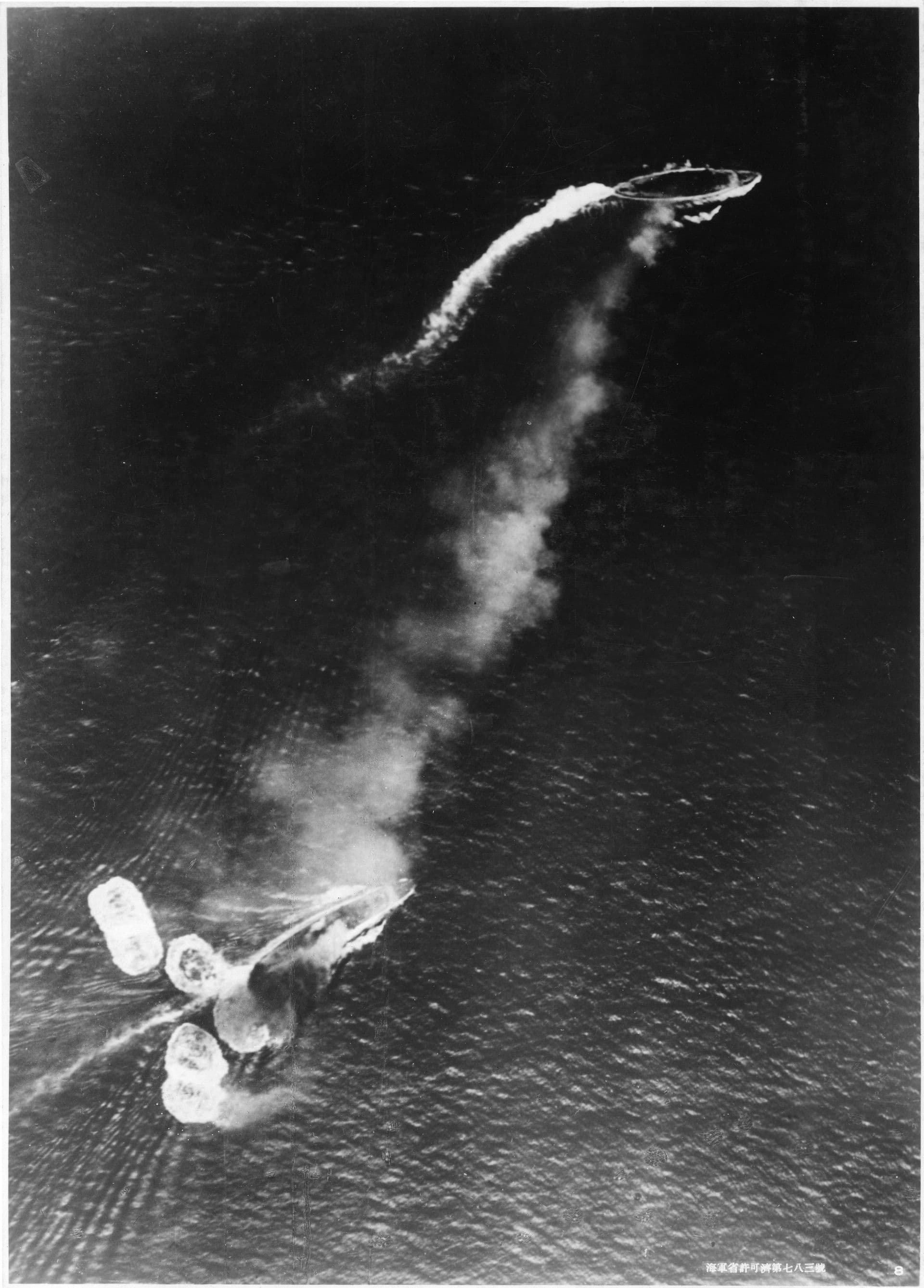 Black‑and‑white aerial photograph showing two large naval ships under attack at sea. One vessel trails thick smoke and is sharply turning, leaving a curved wake across the water, while the second ship steams ahead at speed with a long white wake behind it. Several bomb splashes erupt around the nearer ship, sending plumes of water into the air. The scene captures the moment of a high‑level bombing attack, with the dark ocean stretching out beneath both ships.