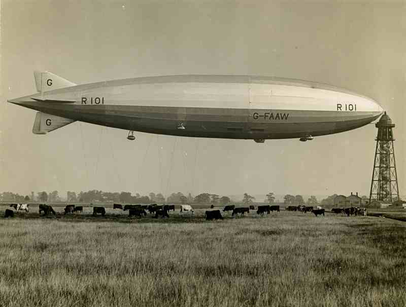 A black‑and‑white photograph of the airship R101 hovering low above an open field. The long, streamlined hull displays the markings “R101” and “G‑FAAW” along its side, with stabilising fins visible at the tail. Below, a herd of cattle grazes in the grassy paddock. At the right of the image stands a tall metal mooring mast, and beyond the field a line of trees and small buildings can be seen under a hazy sky.